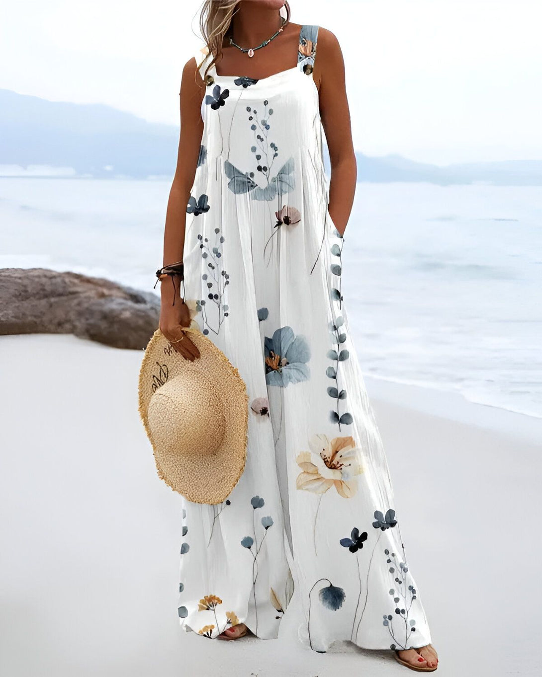 Une femme porte une combinaison pantalon blanche à motifs floraux bleus et verts. Elle est debout sur la plage et tient un chapeau de paille à la main.
