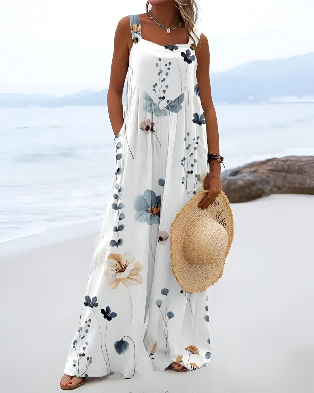 Une femme porte une combinaison-pantalon longue et ample à motifs floraux. Elle est dos à la mer et porte un chapeau de paille à la main.