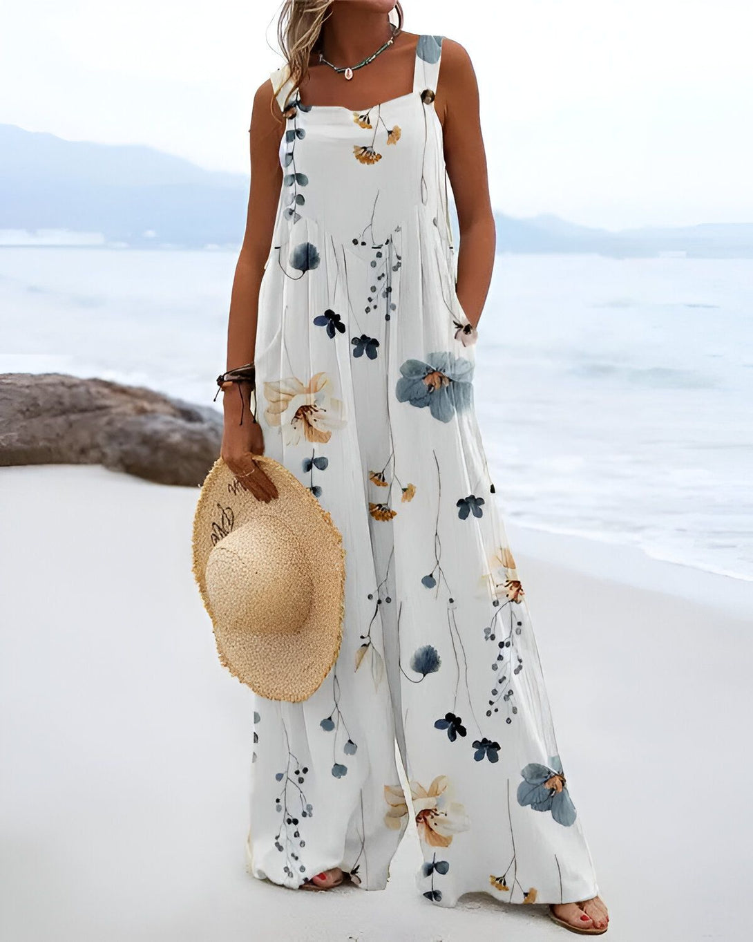 Une femme porte une combinaison pantalon blanche à motifs floraux bleus et jaunes. Elle est debout sur la plage et tient un chapeau de paille à la main.