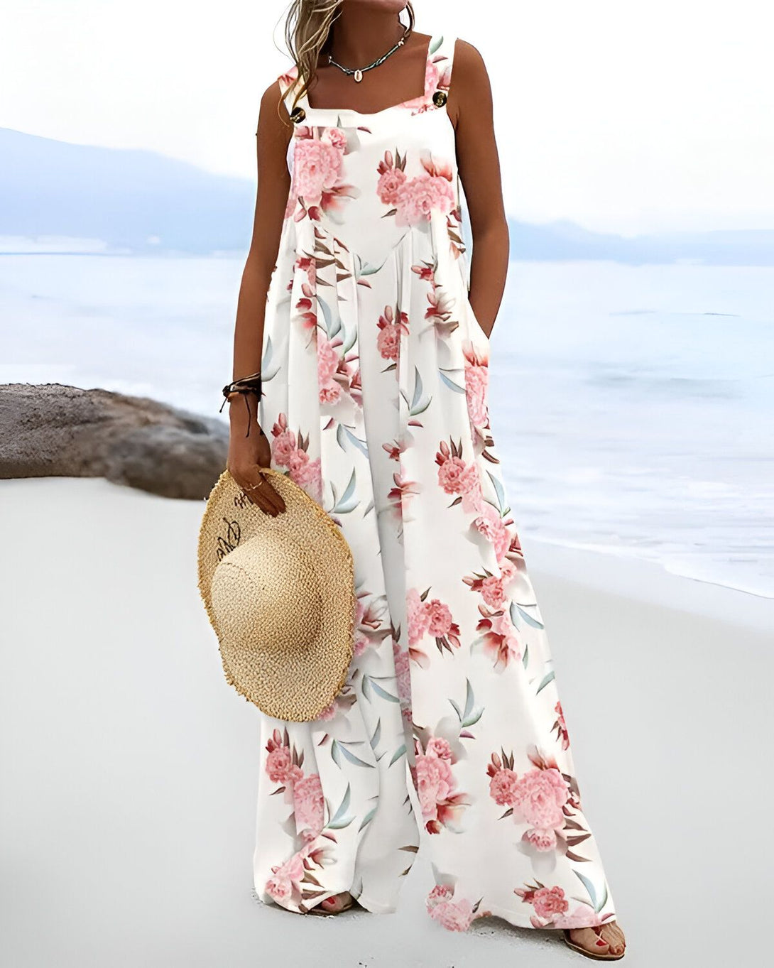 Une femme porte une combinaison pantalon ample à motifs floraux blancs et roses. Elle est dos à la caméra et se tient sur une plage, un chapeau de paille à la main.