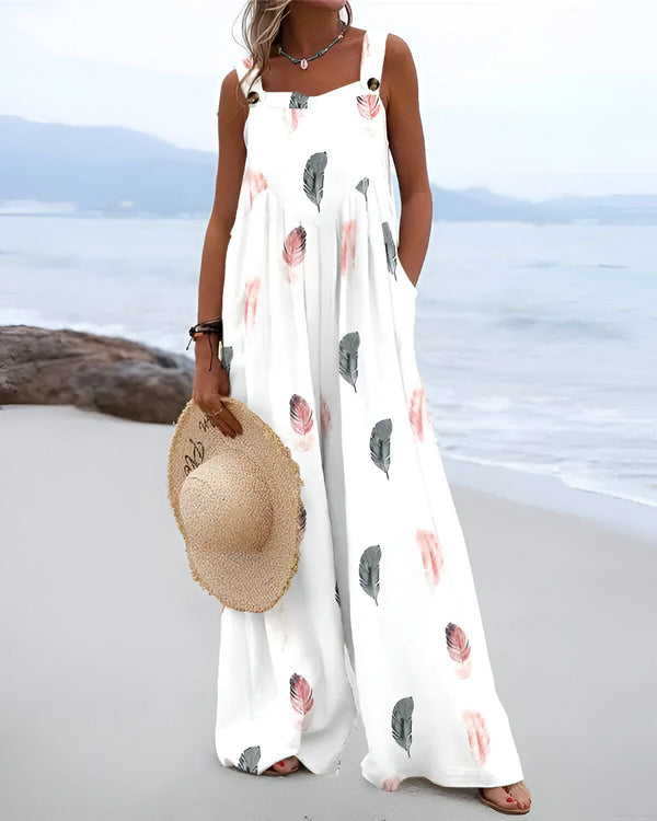Une femme porte une combinaison blanche à motifs de plumes avec un chapeau de paille à la main et se trouve sur la plage.