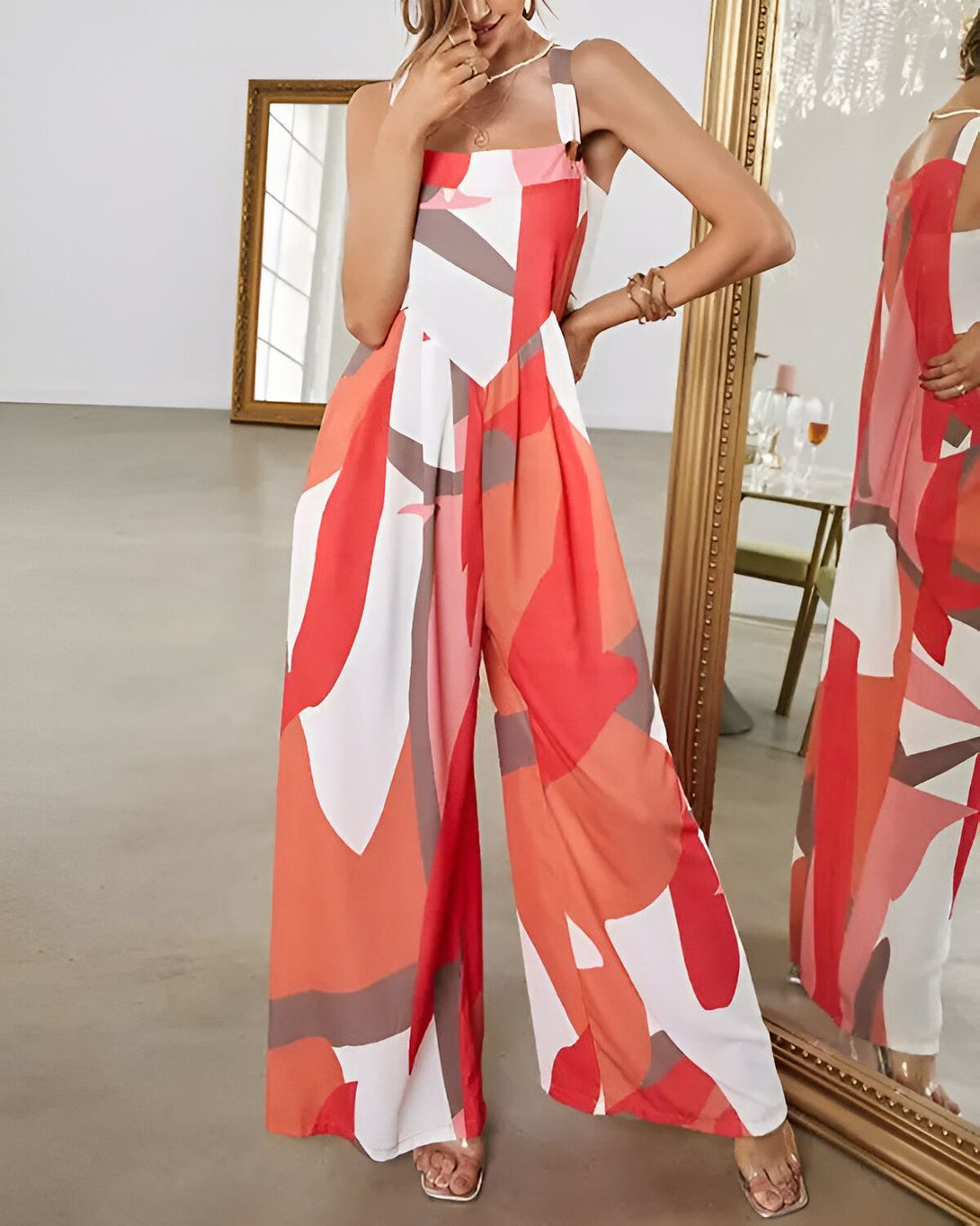 Une femme porte une combinaison pantalon large et coloré. La combinaison est rouge, orange et blanche. Elle a des bretelles fines et un col carré. La femme est debout dans un salon.