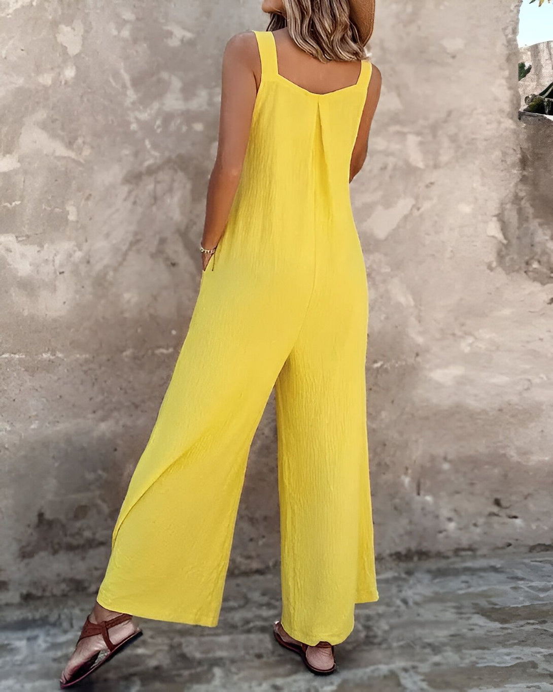 Une femme porte une combinaison-pantalon jaune. Elle est vue de dos. La combinaison-pantalon est ample et a des jambes larges. La femme porte également un chapeau de paille et des sandales marron.