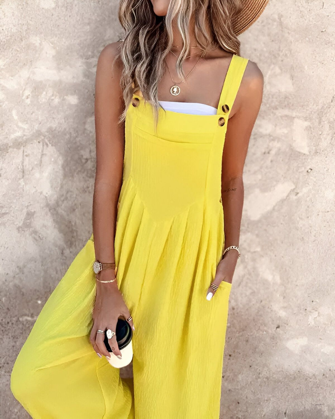 Une femme porte une combinaison-pantalon jaune avec un chapeau de paille et tient une tasse à café à la main.