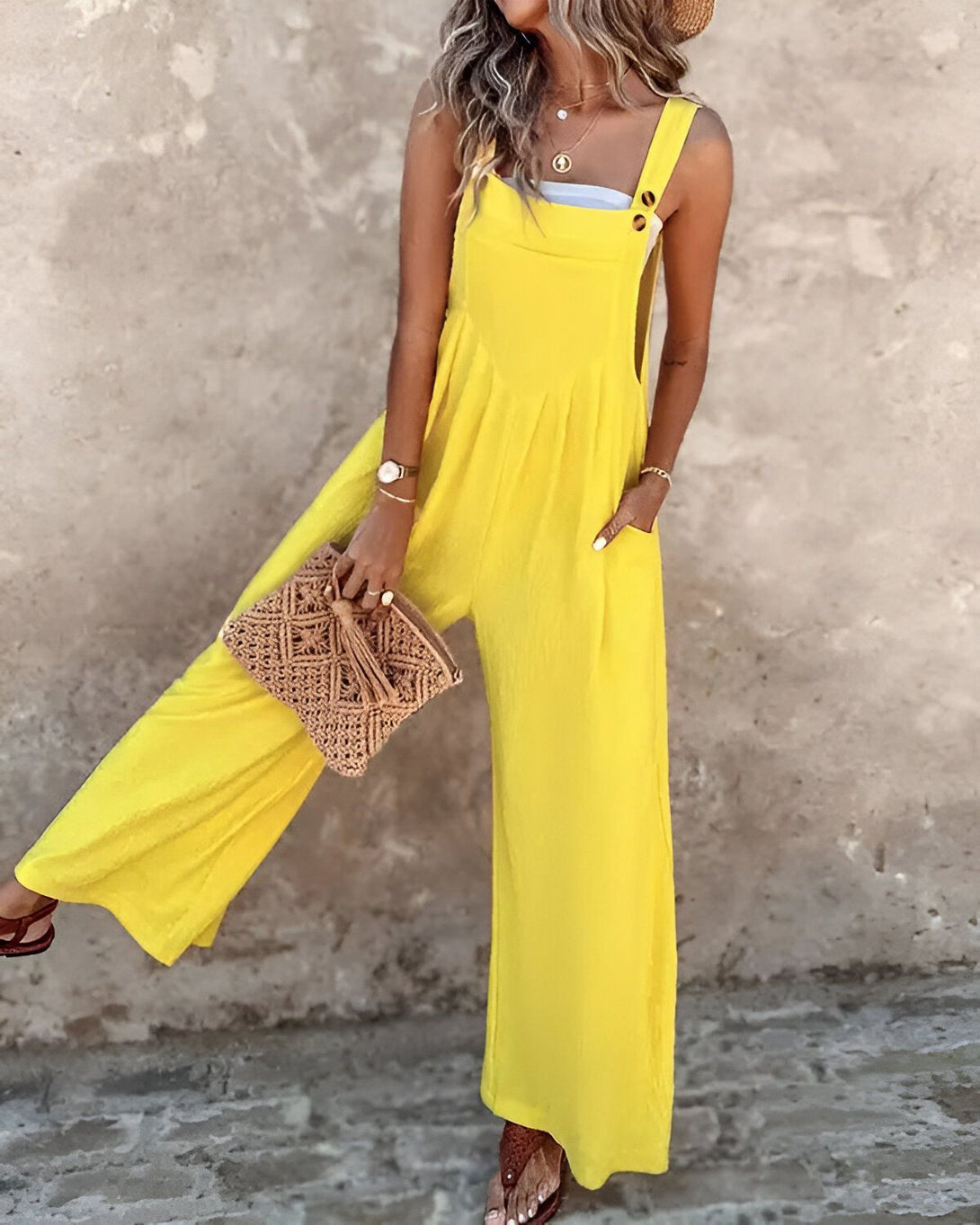 Une femme porte une combinaison-pantalon jaune avec un chapeau de paille et des sandales marron. Elle porte un sac à main en osier marron et un collier.