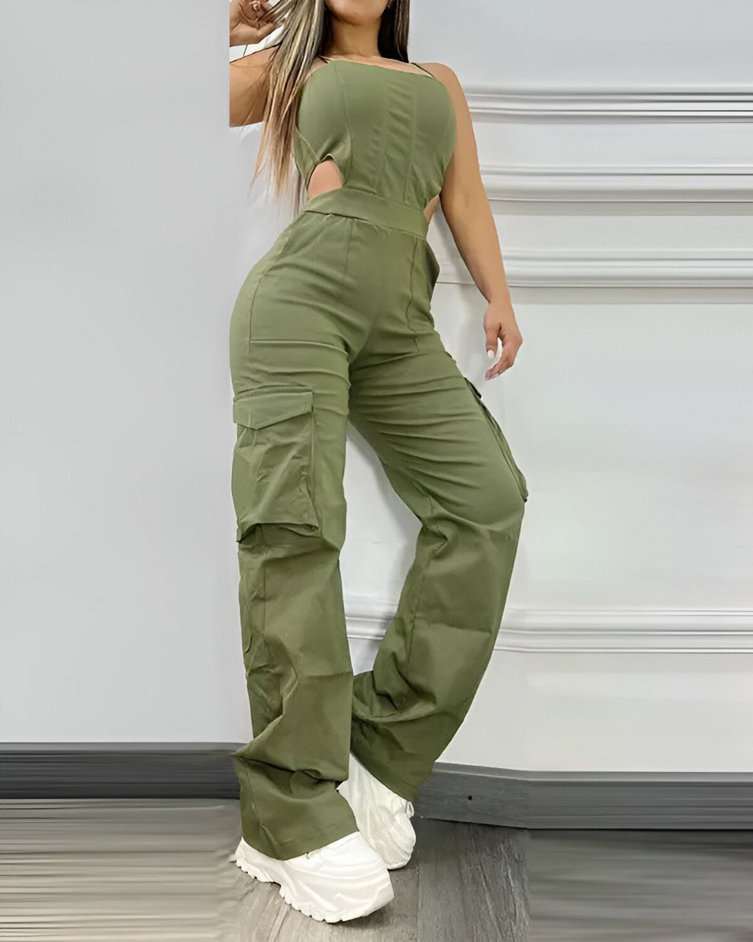 Une femme porte une combinaison-pantalon cargo vert olive. La combinaison est ouverte au niveau de la poitrine et possède des poches cargo sur les côtés. Elle porte des baskets blanches.
