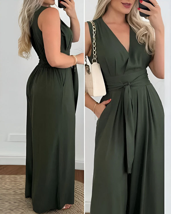 Une femme porte une combinaison-pantalon verte. La combinaison-pantalon est sans manches et possède une ceinture à la taille. La femme est blonde et porte un sac à main blanc.