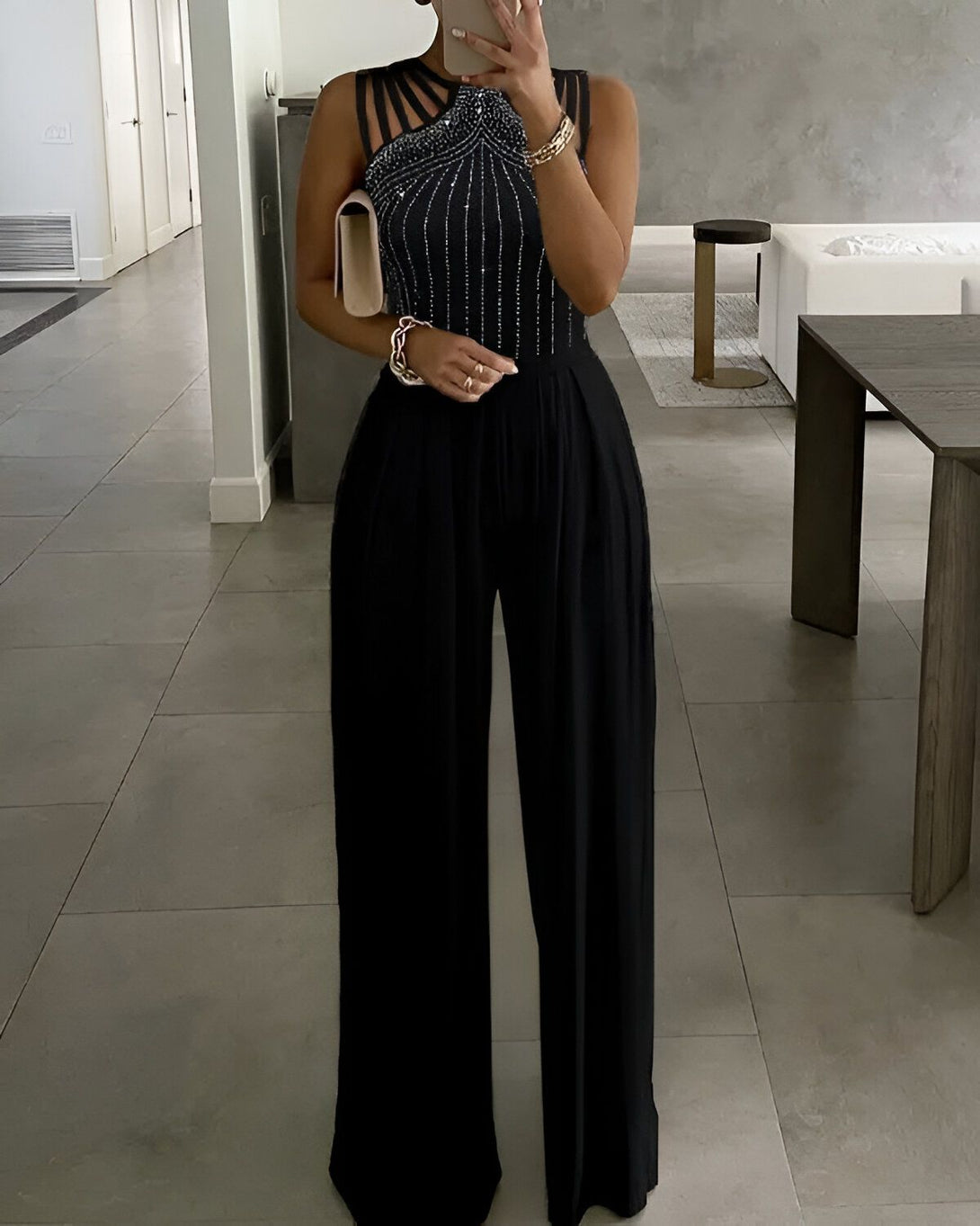 Une femme porte une combinaison pantalon noire avec des sandales à talons noires. Elle a un sac à main beige à la main.