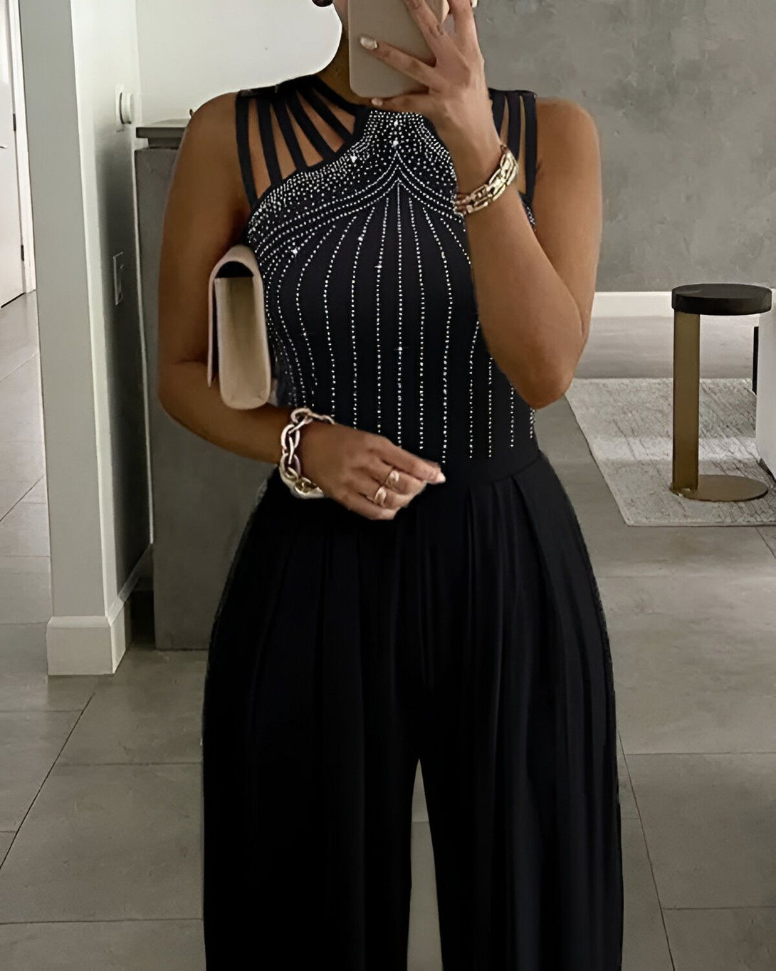 Une femme porte une combinaison noire avec des strass. Elle a les cheveux longs et noirs et porte un maquillage glamour. Elle est debout dans un couloir et tient un sac à main beige.