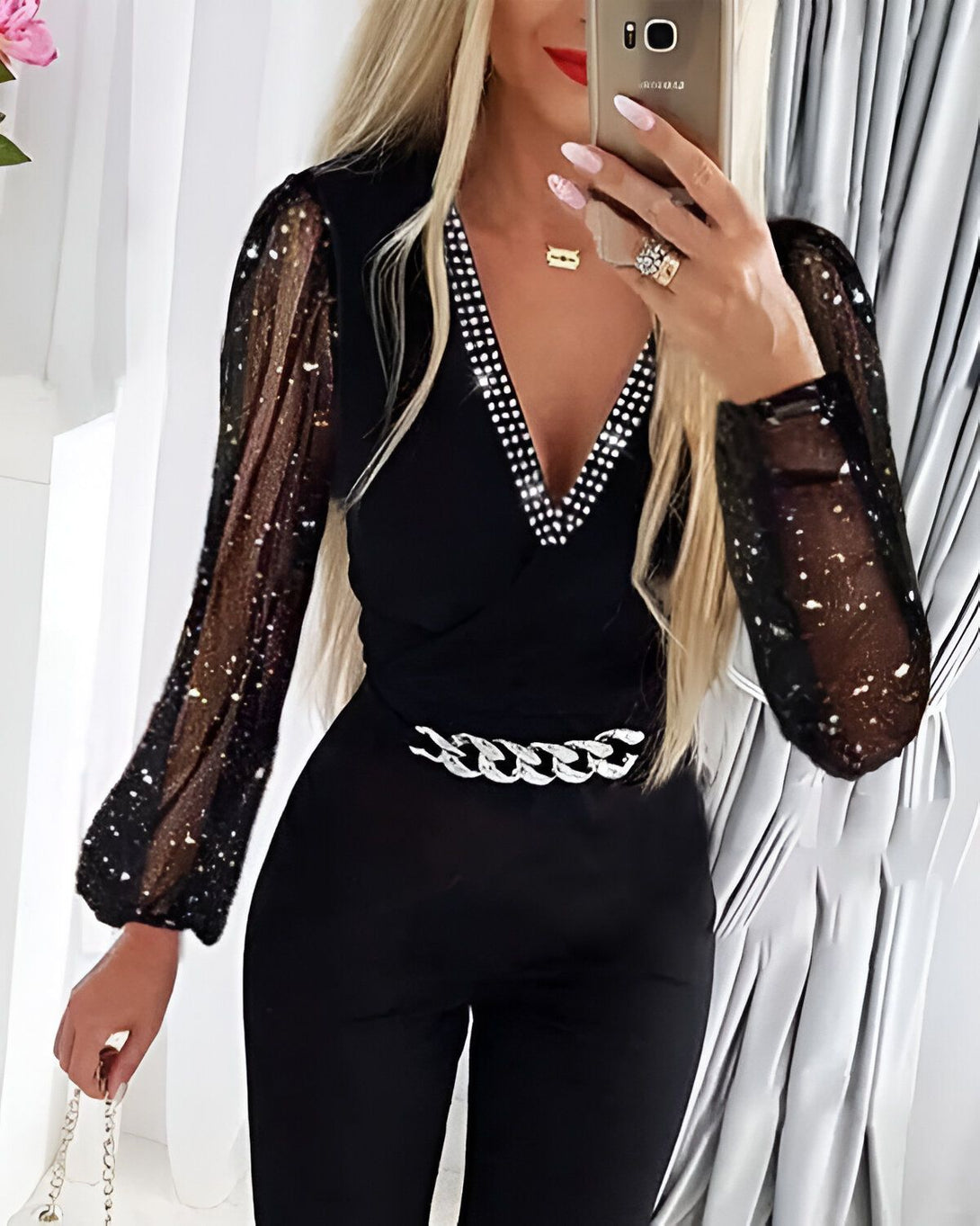 Une femme porte une combinaison noire avec un décolleté en V et des manches longues bouffantes. Elle porte également une ceinture à chaîne argentée et un collier.