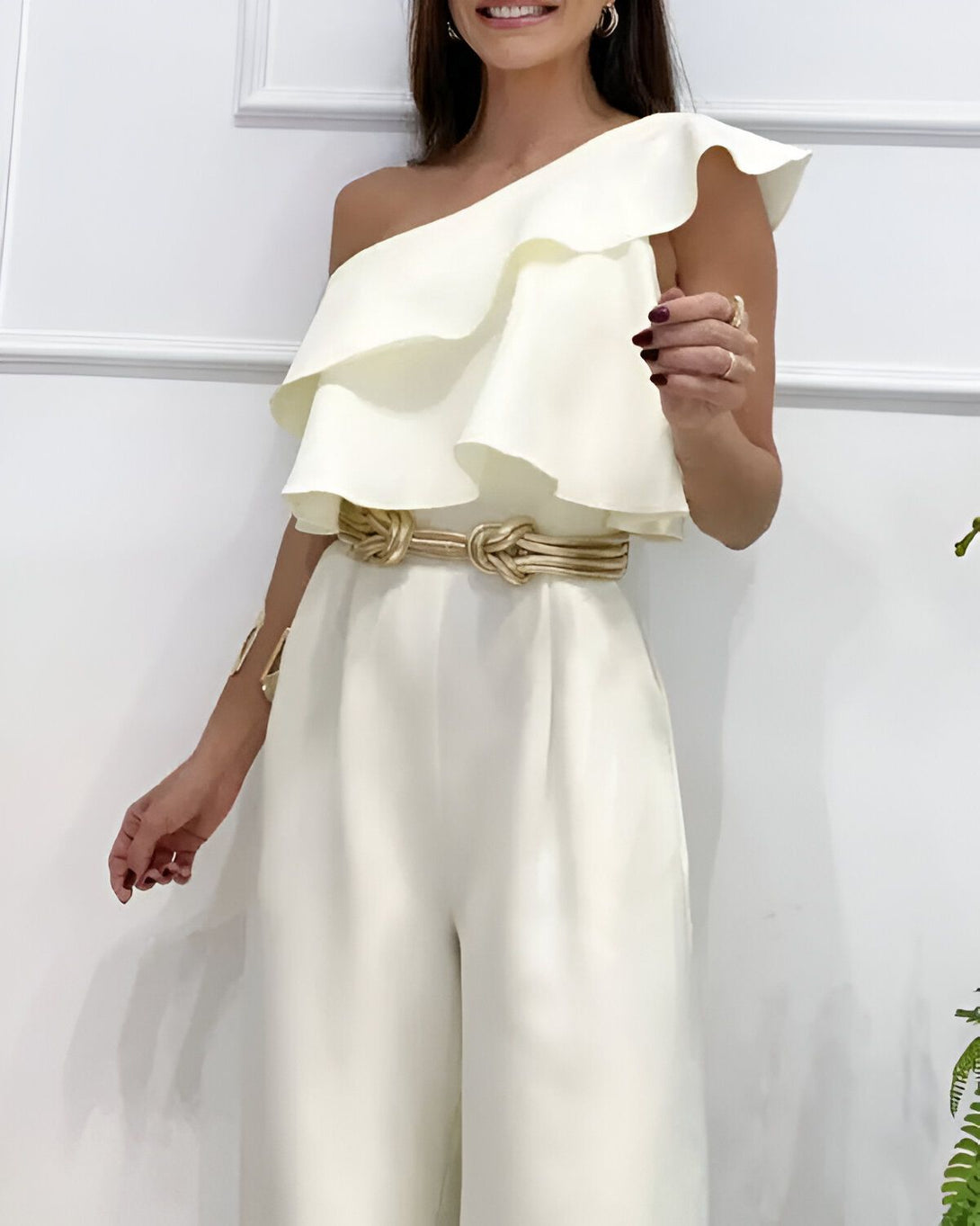 Une femme porte une combinaison blanche à une épaule avec une ceinture dorée.