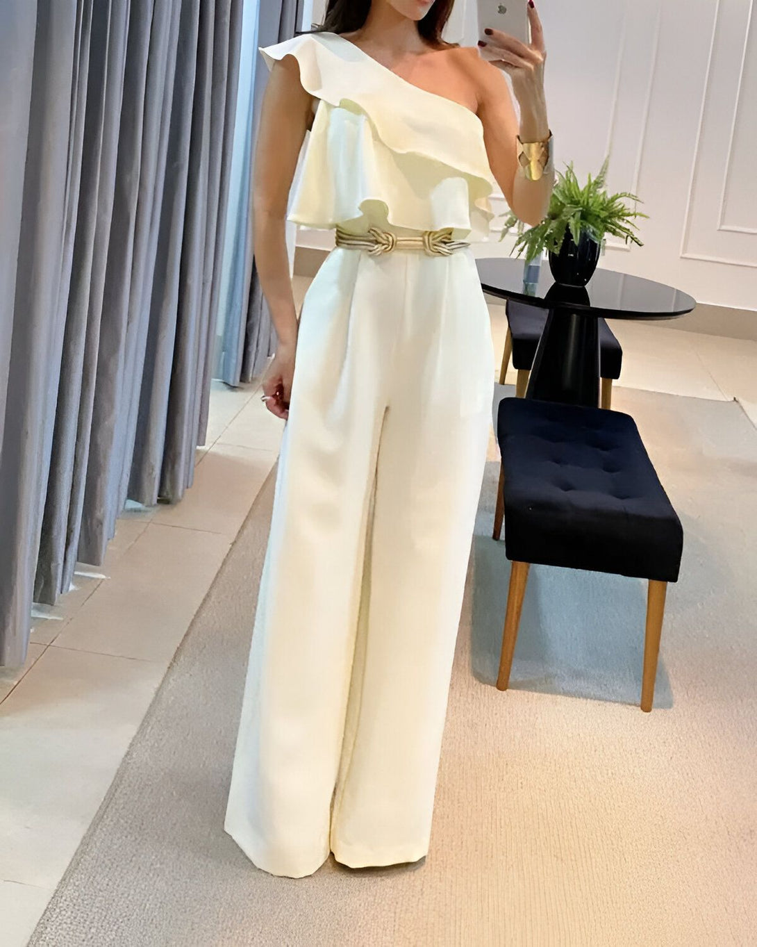 Une femme porte une combinaison pantalon blanche avec une ceinture dorée. Elle a une manche bouffante et l'autre manche est nue. Elle porte des talons hauts blancs et un bracelet doré.