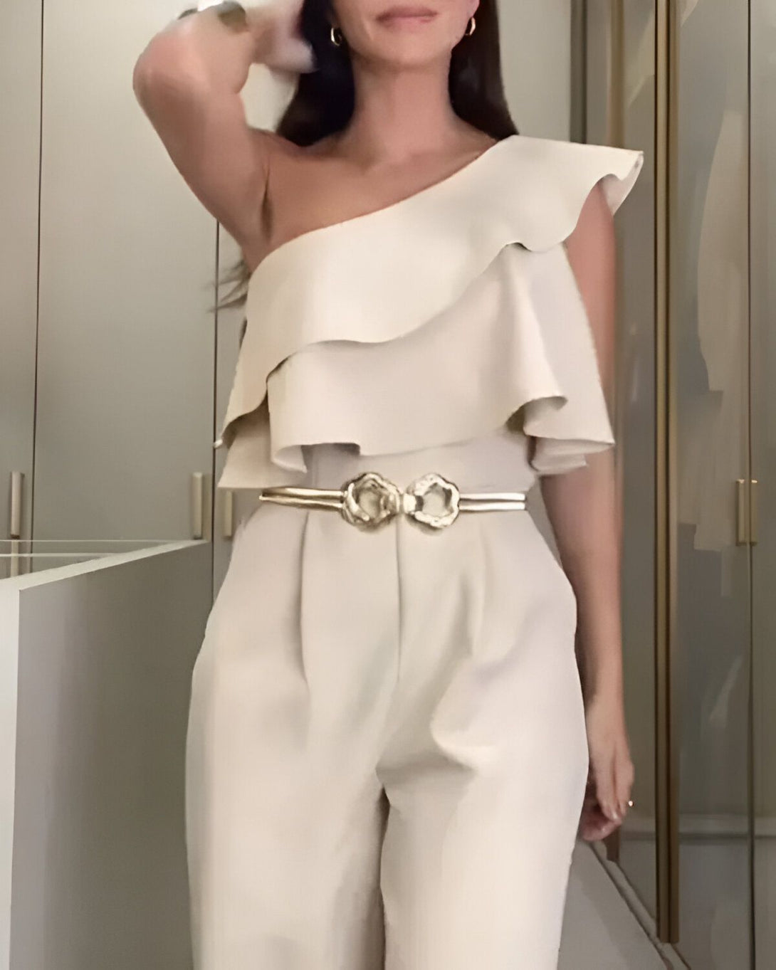 Une femme porte une combinaison pantalon beige avec une manche et une jambe. Elle est ceinturée d'une ceinture dorée.