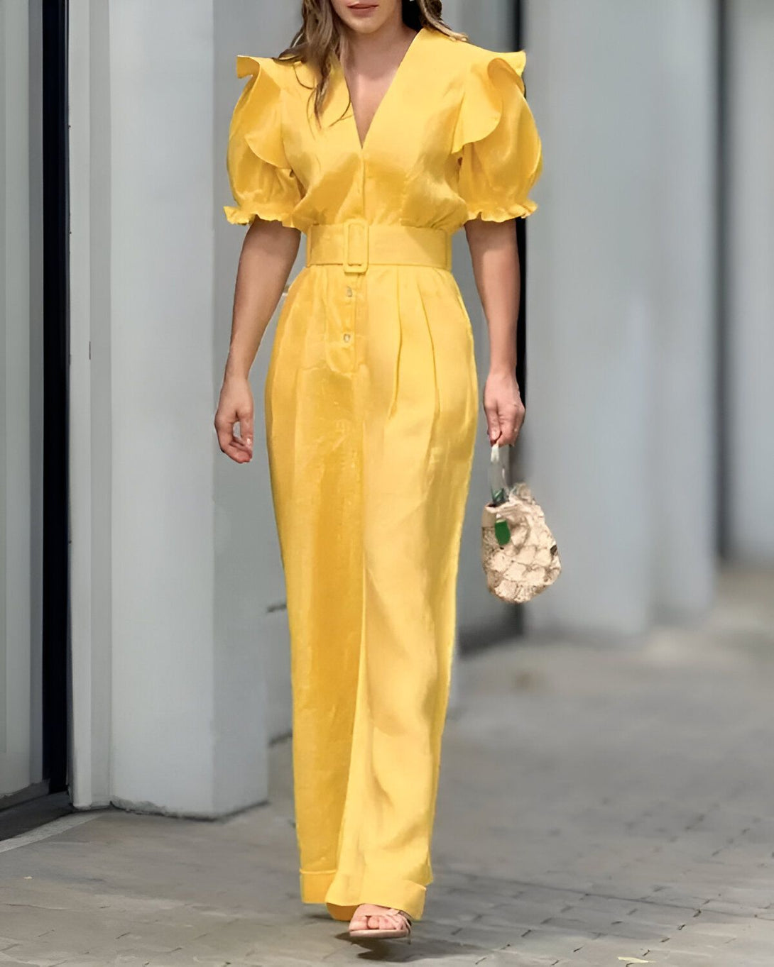 Une femme porte une combinaison-pantalon jaune avec des manches bouffantes et un pantalon large. Elle est ceinturée à la taille et porte des sandales à talons hauts. Elle a un sac à main en osier à la main.