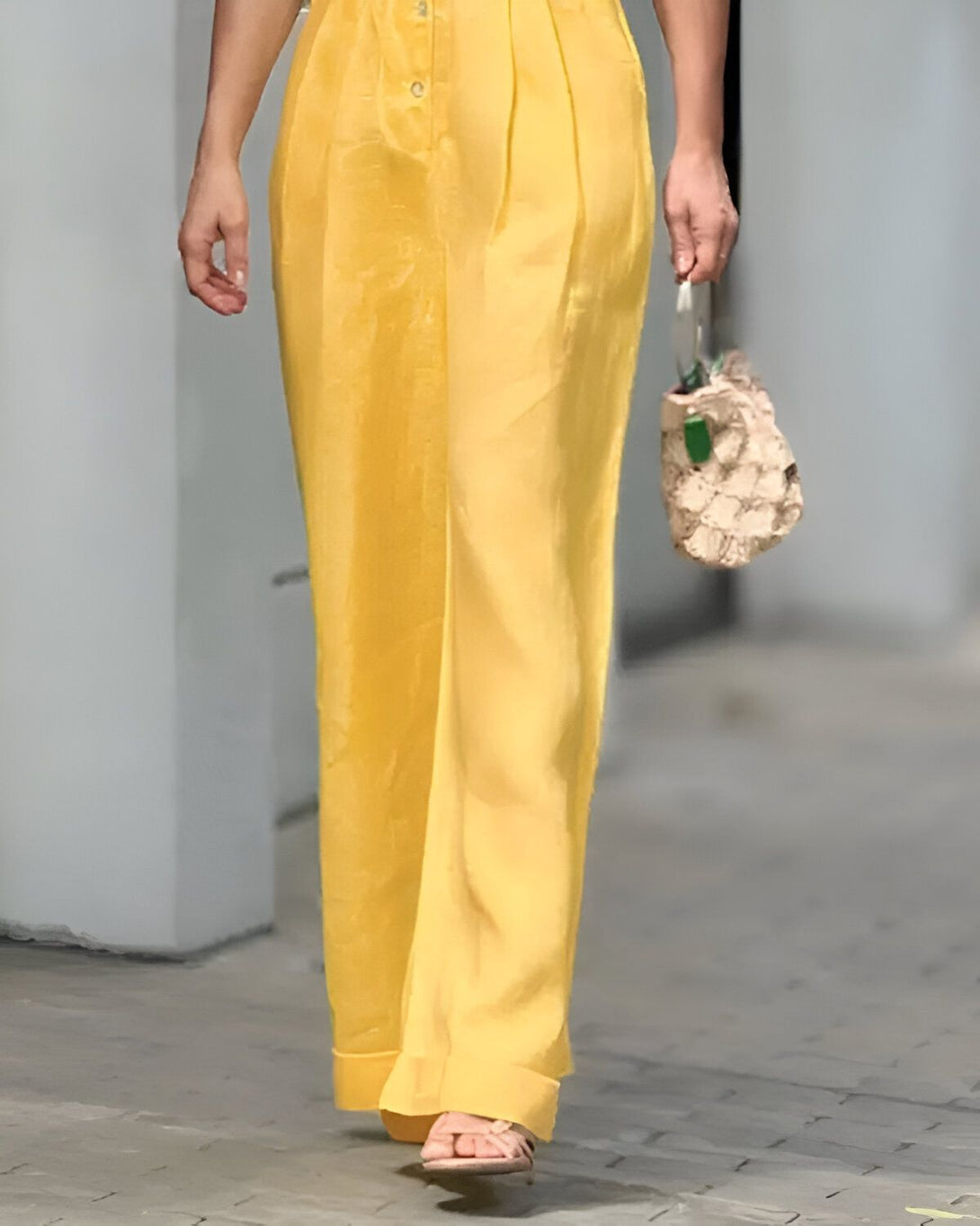 Une femme porte un pantalon jaune large et une sandale marron. Elle porte un sac à main beige et marron.
