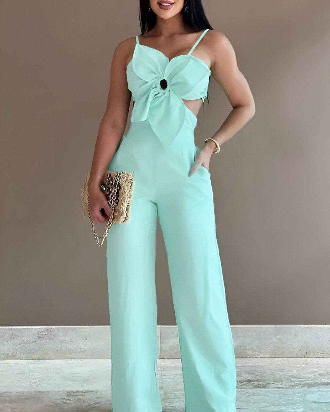 Une femme porte une combinaison pantalon vert menthe. La combinaison a un haut court avec un nœud papillon sur le devant et des jambes larges. La femme porte également un sac à main en paille et des talons hauts.
