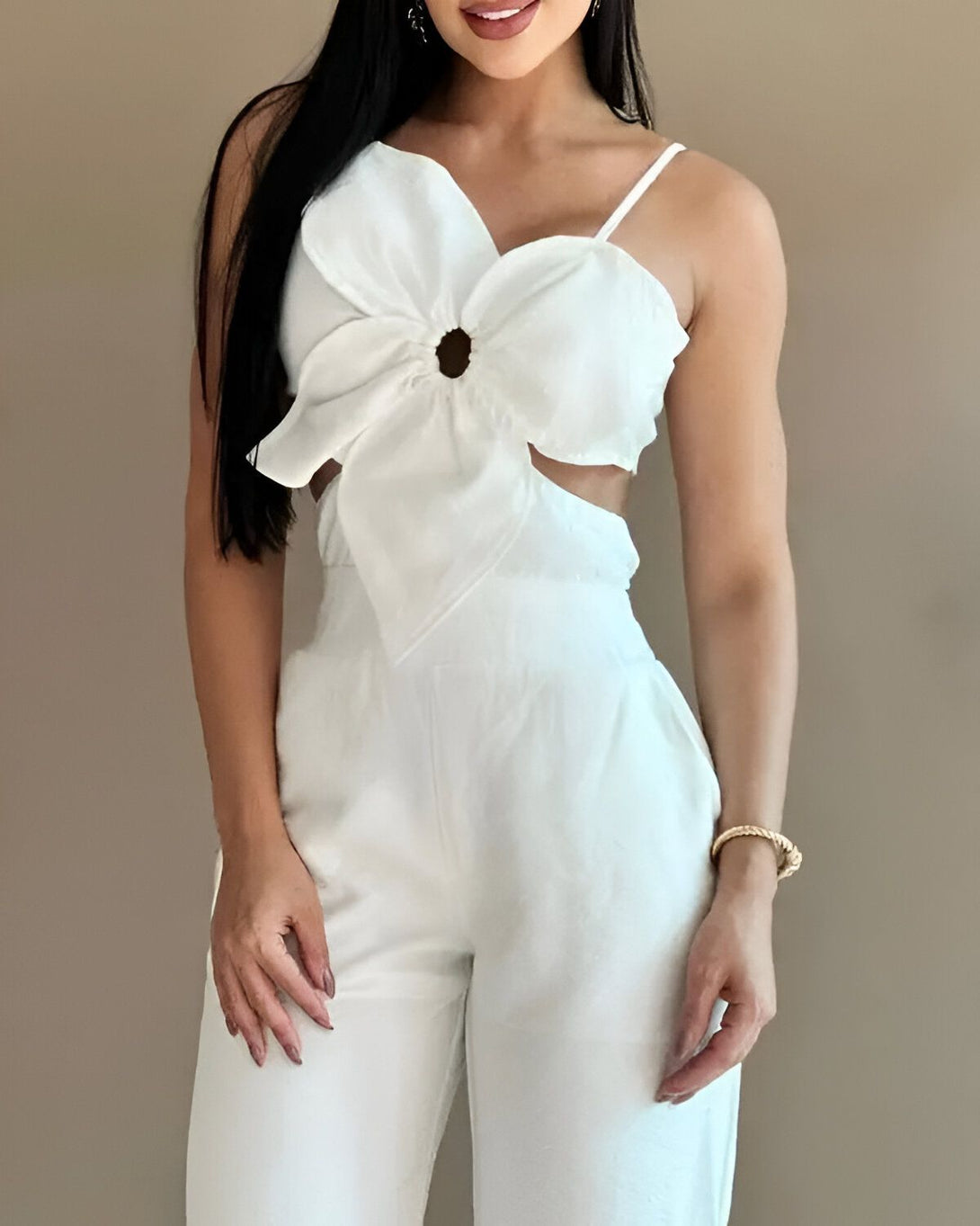 Une femme porte une combinaison pantalon blanche. La combinaison a un haut court avec un grand nœud sur le devant. Le pantalon est large et évasé. La femme est souriante et porte des talons hauts blancs.