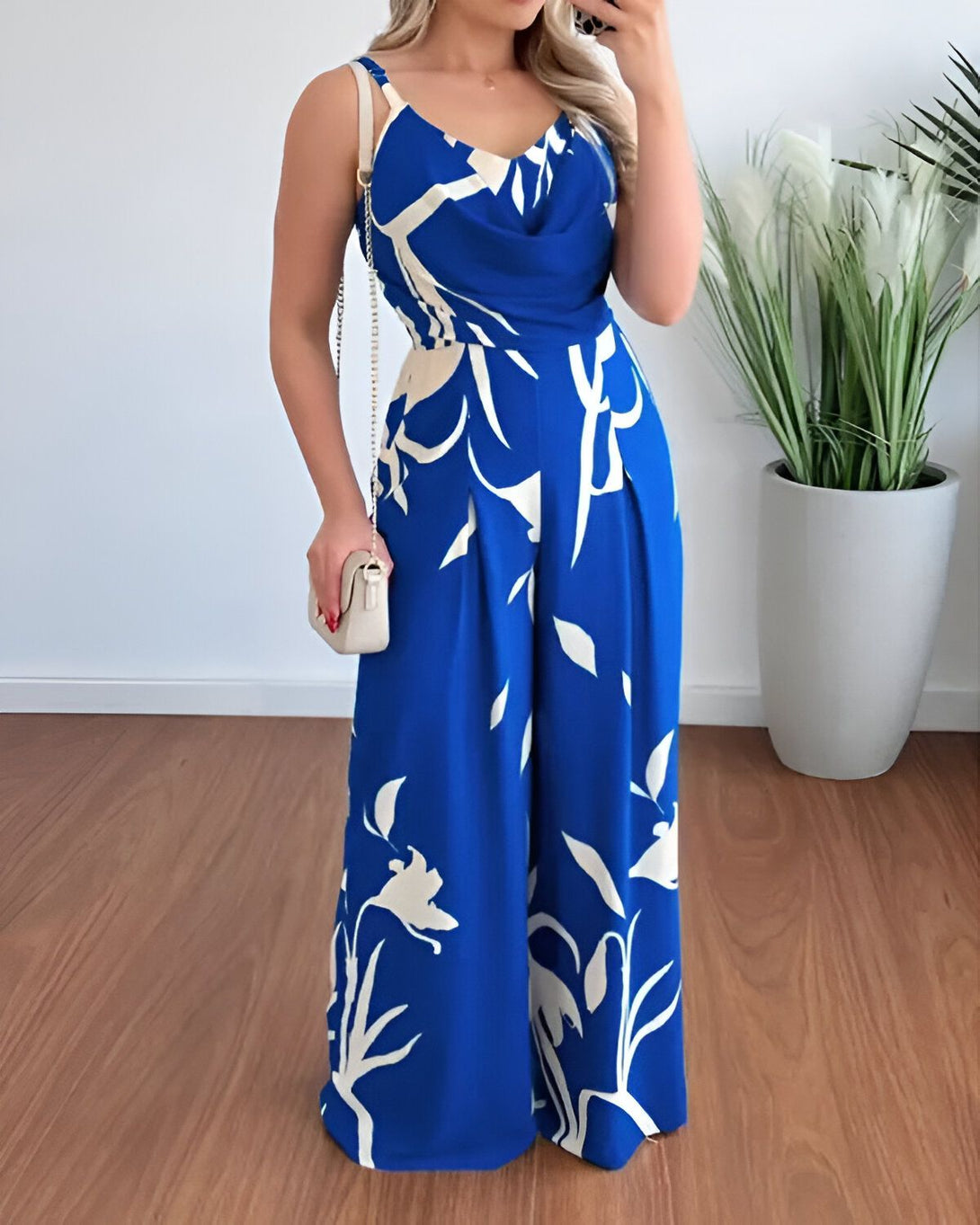 Une femme porte une combinaison pantalon bleue à motifs blancs. Elle est dos nu et a un décolleté plongeant. La combinaison est cintrée à la taille et évasée sur les jambes. La femme porte des sandales à talons hauts et un sac à main blanc.