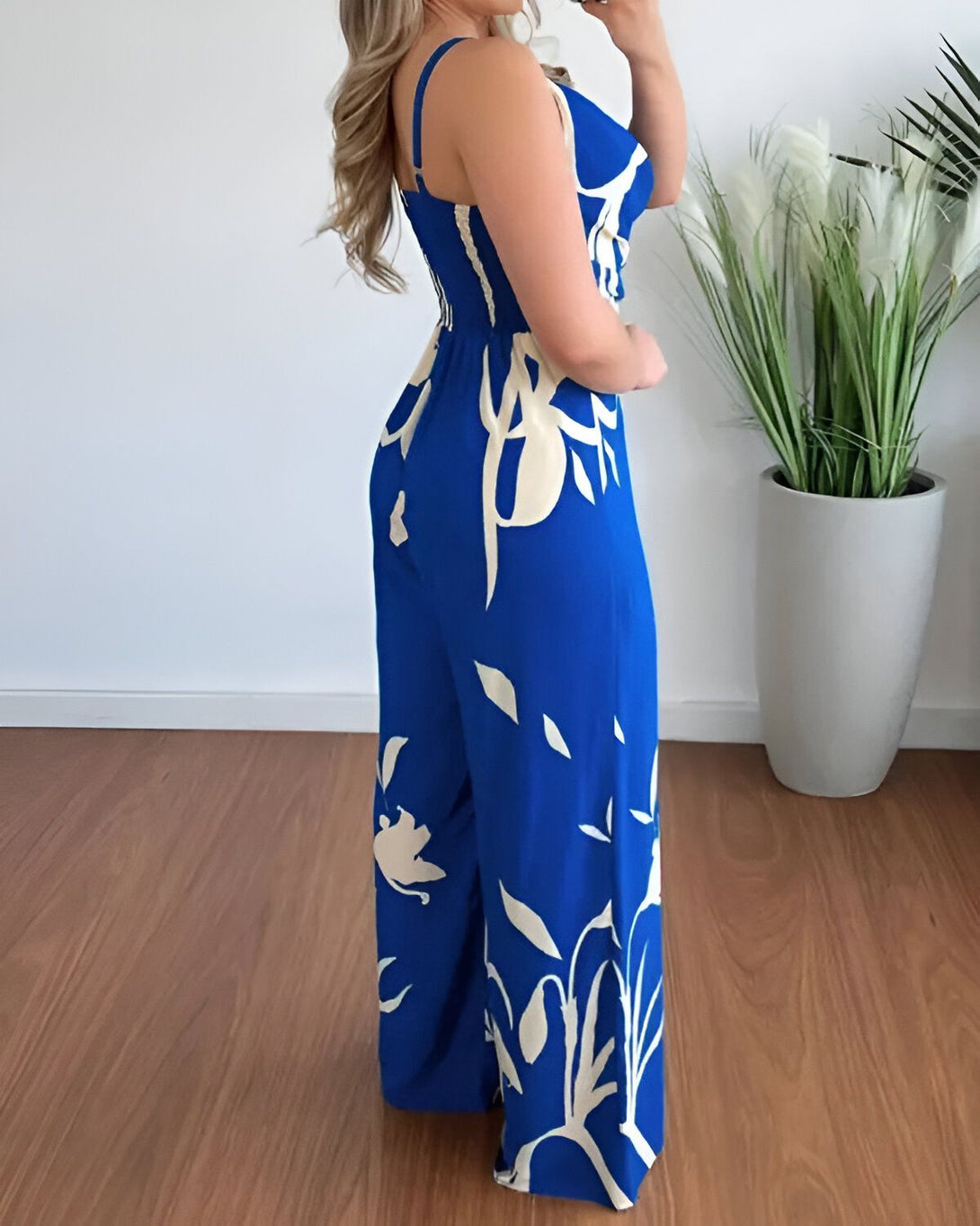 Une femme porte une combinaison pantalon bleue à motifs blancs. La combinaison est cintrée à la taille et possède des jambes larges. La femme est blonde et porte des talons hauts blancs. Elle est debout dans un salon, à côté d'une plante verte.