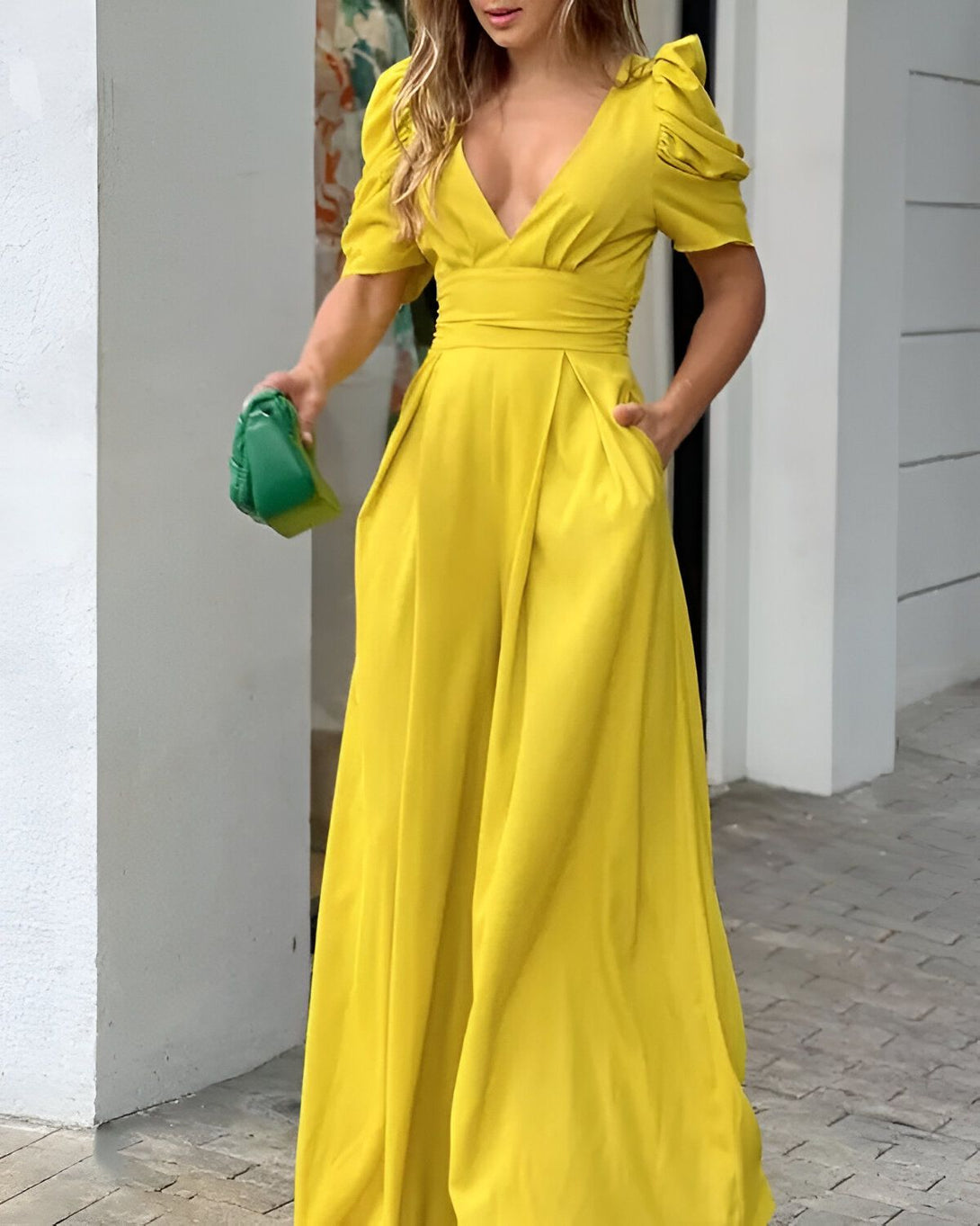 Une femme porte une combinaison-pantalon jaune vif. Elle a les cheveux longs et blonds, et elle porte un sac à main vert. Elle est debout dans une rue, et il y a des bâtiments en arrière-plan.