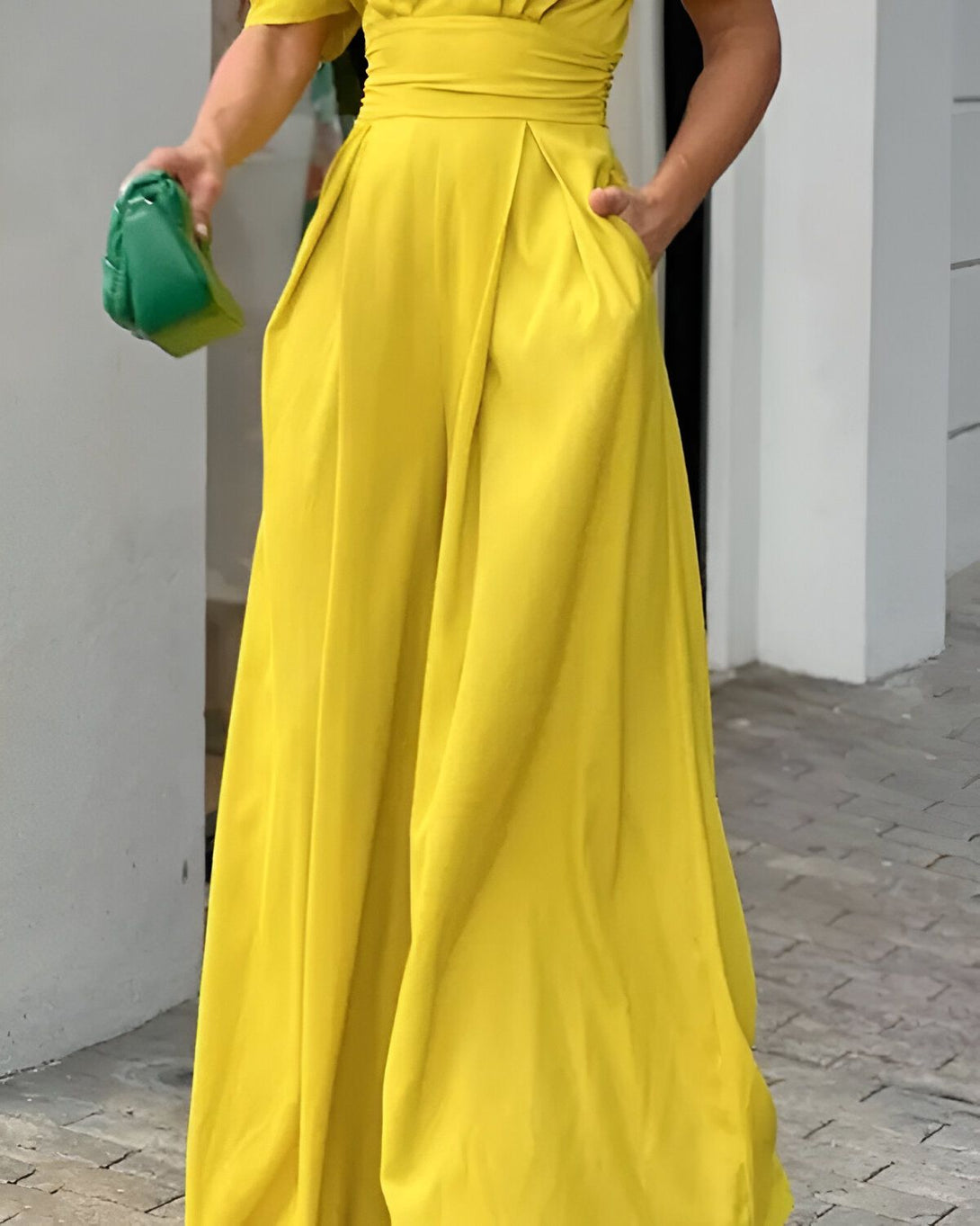 Une femme porte une combinaison-pantalon jaune avec des sandales à talons jaunes. Elle a un sac à main vert à la main.