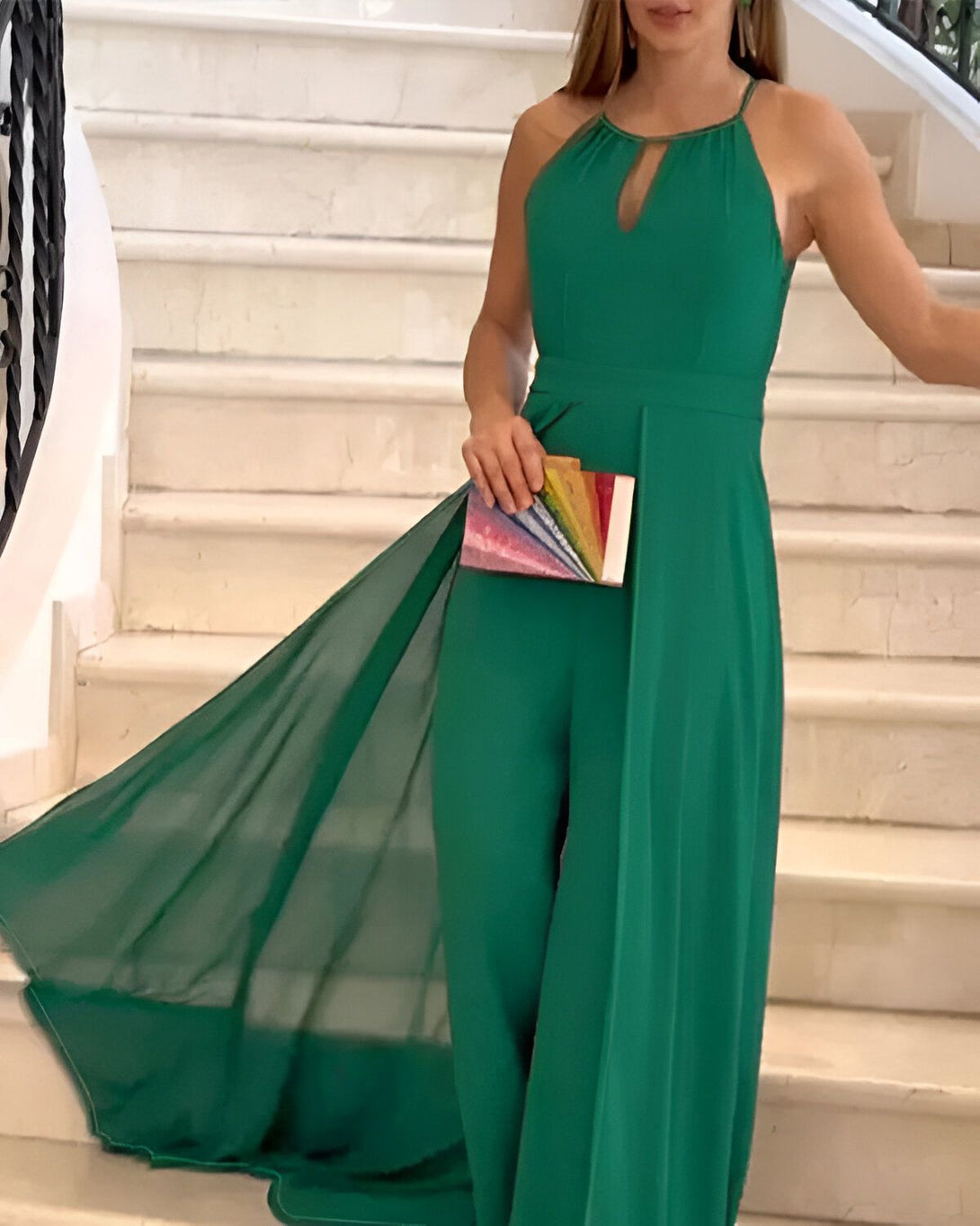 Une femme portant une combinaison-pantalon vert émeraude avec un décolleté plongeant et une jambe évasée. Elle porte également une pochette arc-en-ciel à la main.
