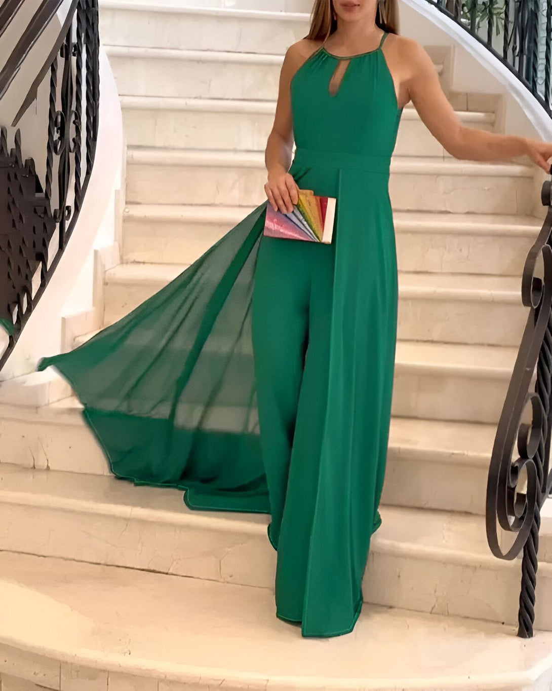 Une femme portant une combinaison-pantalon verte fendue sur le côté avec une traîne en mousseline de soie. Elle porte également des sandales à talons hauts et une pochette arc-en-ciel.