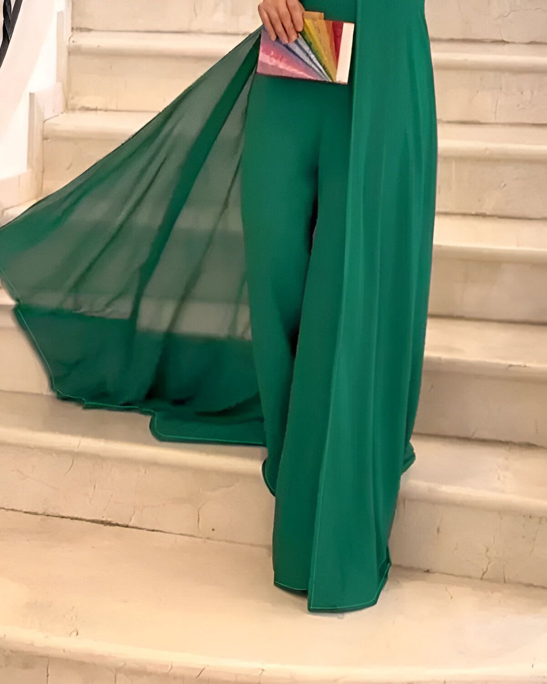 Une femme portant une combinaison pantalon verte et une cape verte transparente. Elle est debout sur unescalier en marbre blanc.