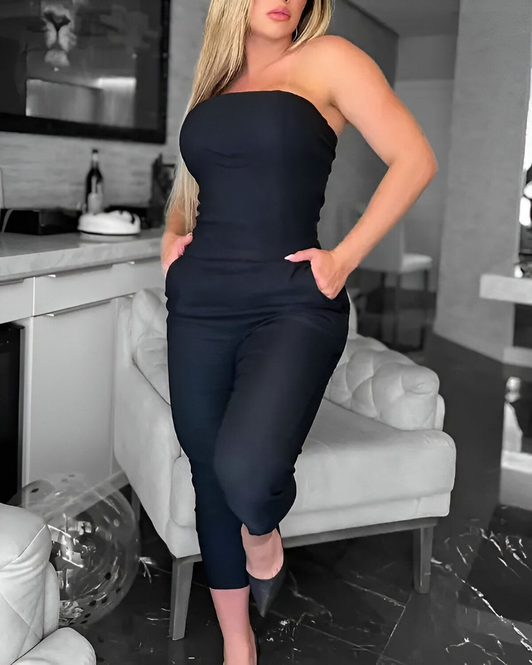 Une femme porte une combinaison-pantalon noire sans bretelles. Elle est debout dans un salon, la main droite dans la poche et la main gauche sur la hanche. Elle porte des talons noirs.