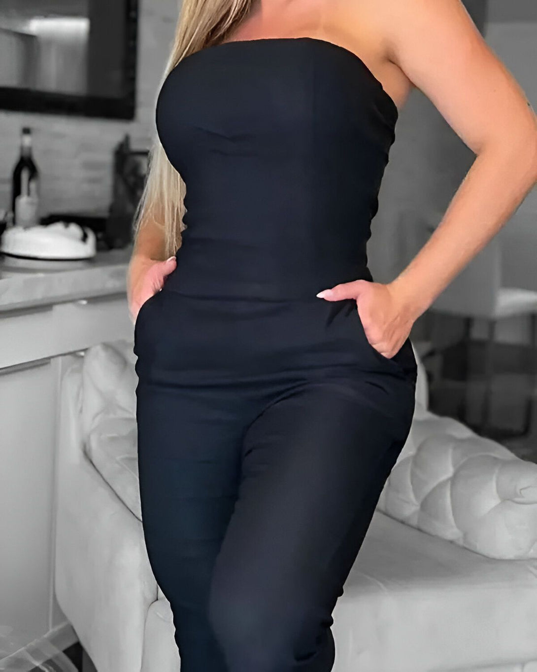 Une femme porte une combinaison noire sans bretelles. Elle a les mains dans les poches et se tient debout dans une pièce.