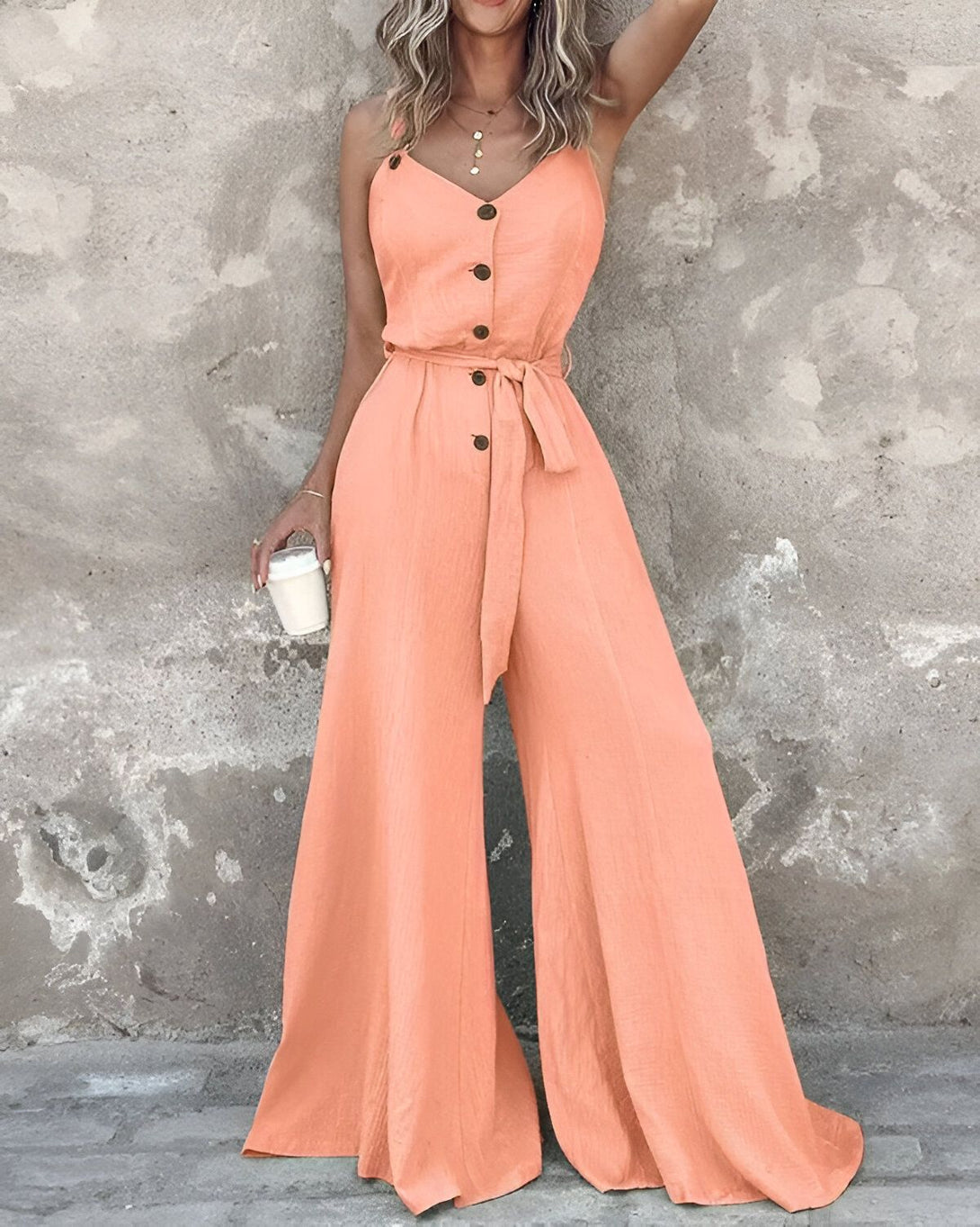 Une femme porte une combinaison-pantalon orange à bretelles. La combinaison est ample et a des jambes larges. Elle est ceinturée à la taille. La femme porte également un collier et un bracelet. Elle a les cheveux longs et blonds et elle est maquillée. Elle est debout devant un mur beige.