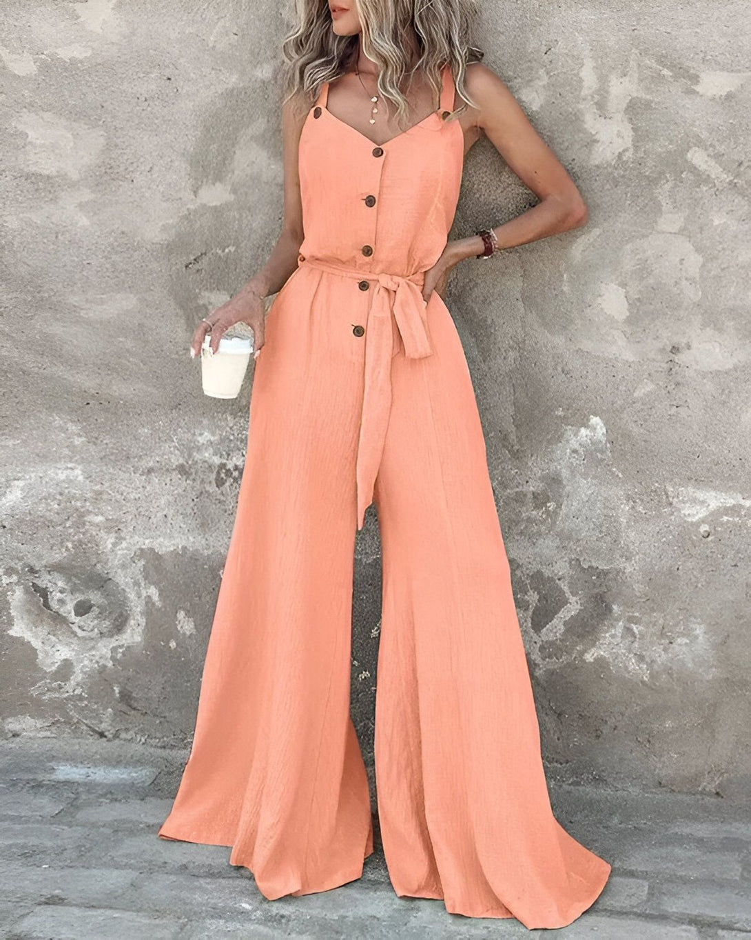 Une femme porte une combinaison-pantalon orange à bretelles. Elle est ceinturée à la taille et porte un café à la main.