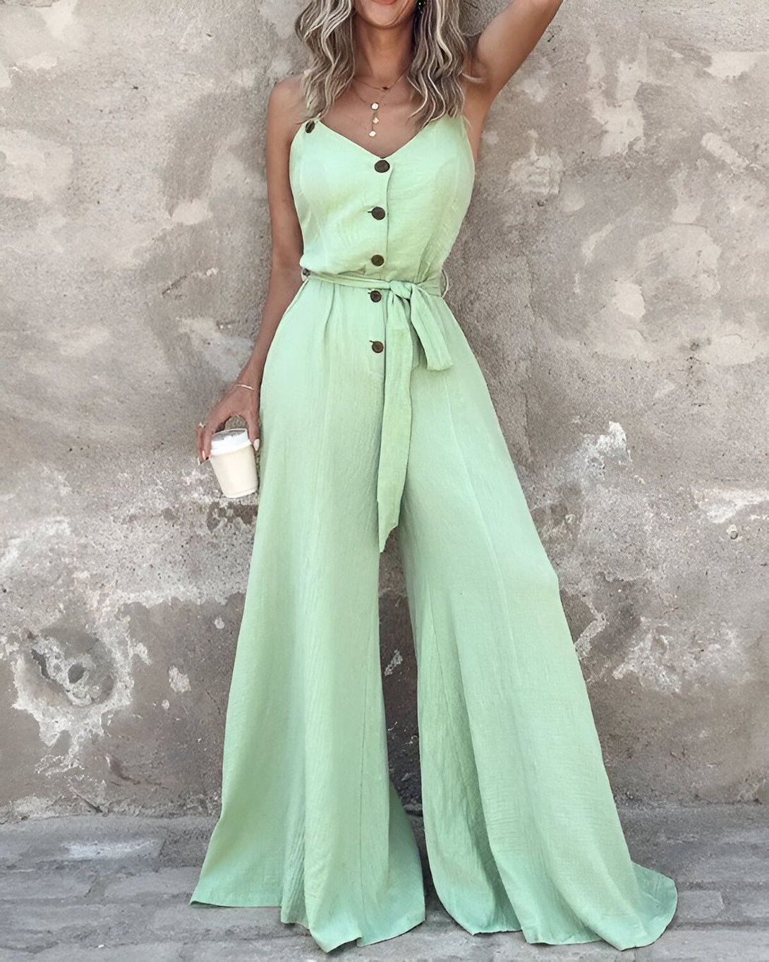 Une femme porte une combinaison-pantalon vert clair. La combinaison a un décolleté en V, des boutons sur le devant et une ceinture à la taille. La femme est debout, un café à la main droite.