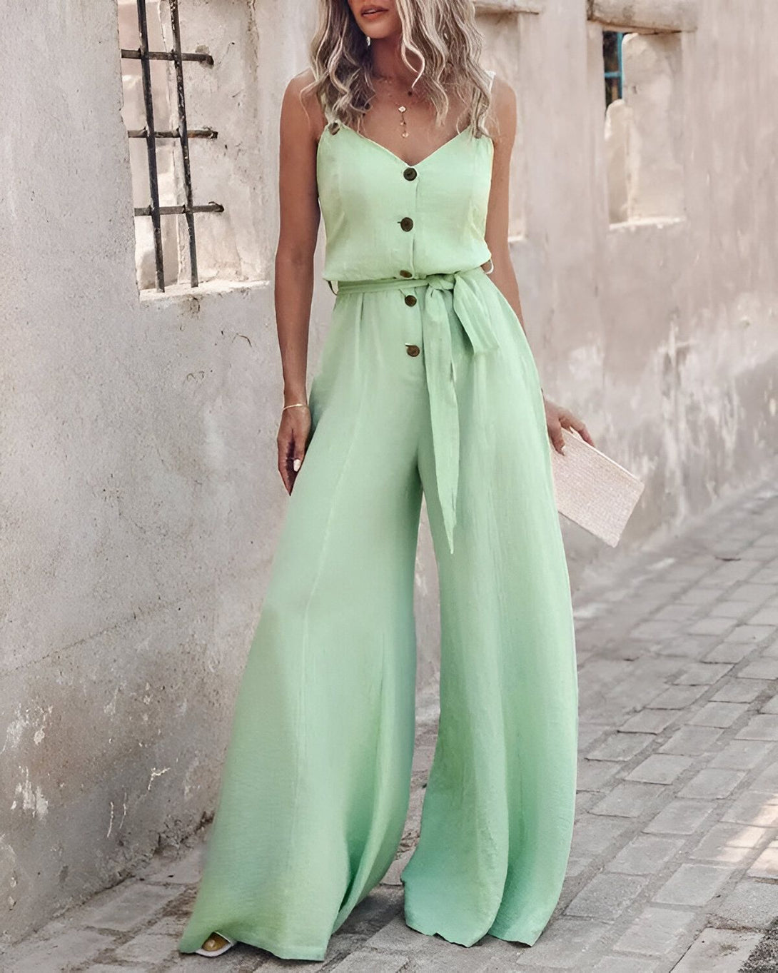 Une femme porte une combinaison-pantalon vert clair. La combinaison a des bretelles fines, un décolleté en V, des boutons sur le devant et une ceinture à la taille. La femme est debout devant un mur et porte des sandales marron clair.