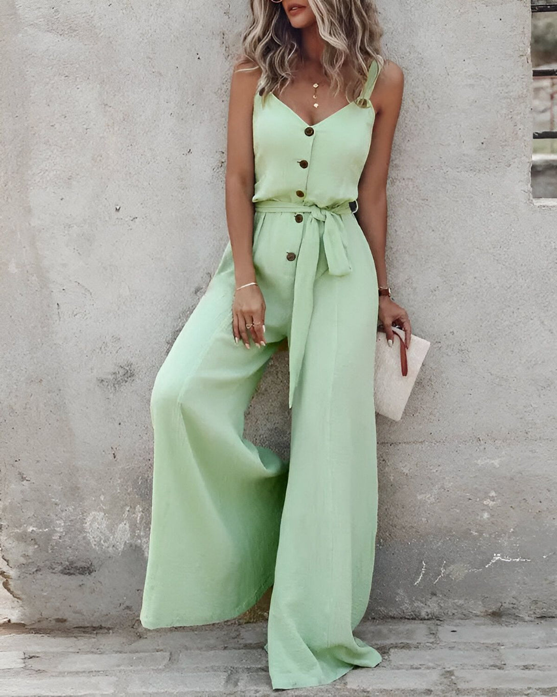 Une femme porte une combinaison-pantalon verte avec un décolleté en V, des boutons et une ceinture à la taille. Elle est associée à des sandales à talons hauts et un sac à main blanc.