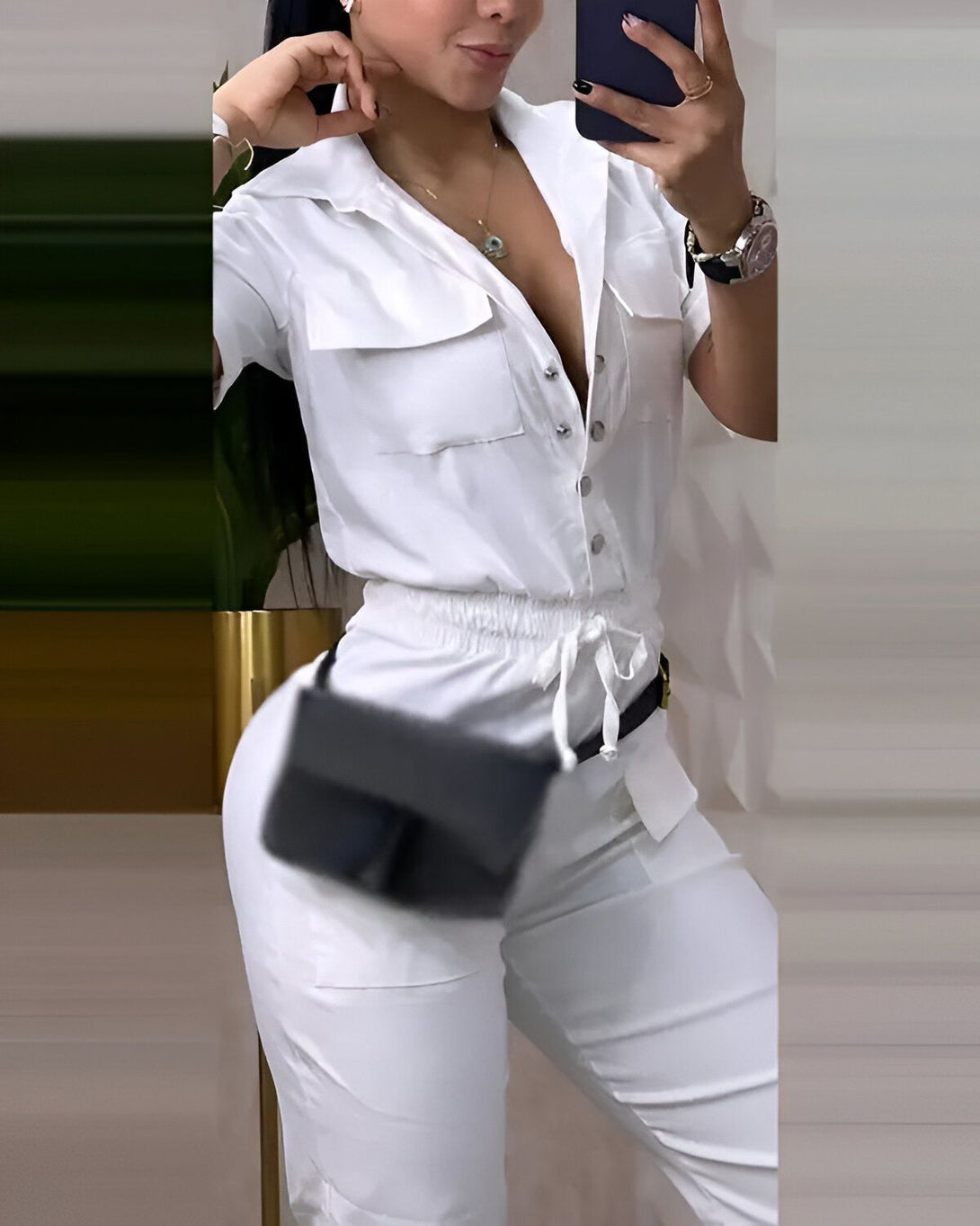 Une femme porte une combinaison blanche avec un décolleté plongeant et des manches courtes. Elle porte également une ceinture noire et un sac à main noir.