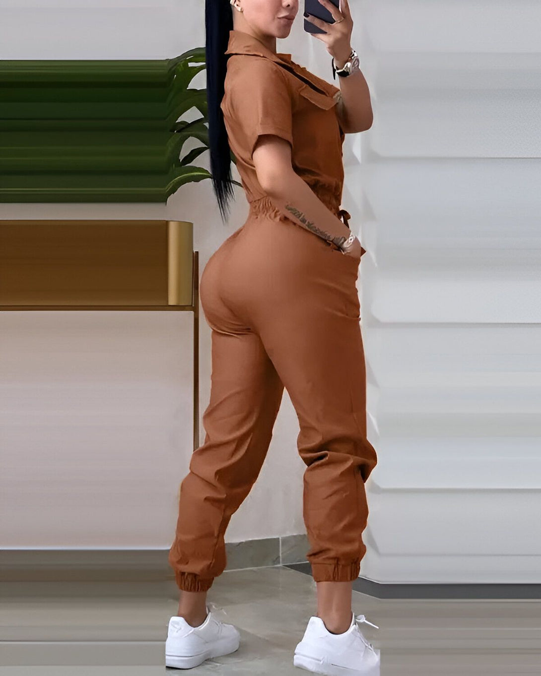 Une femme porte une combinaison marron avec des baskets blanches. Elle est vue de dos et se tient debout, le bras gauche sur la hanche.