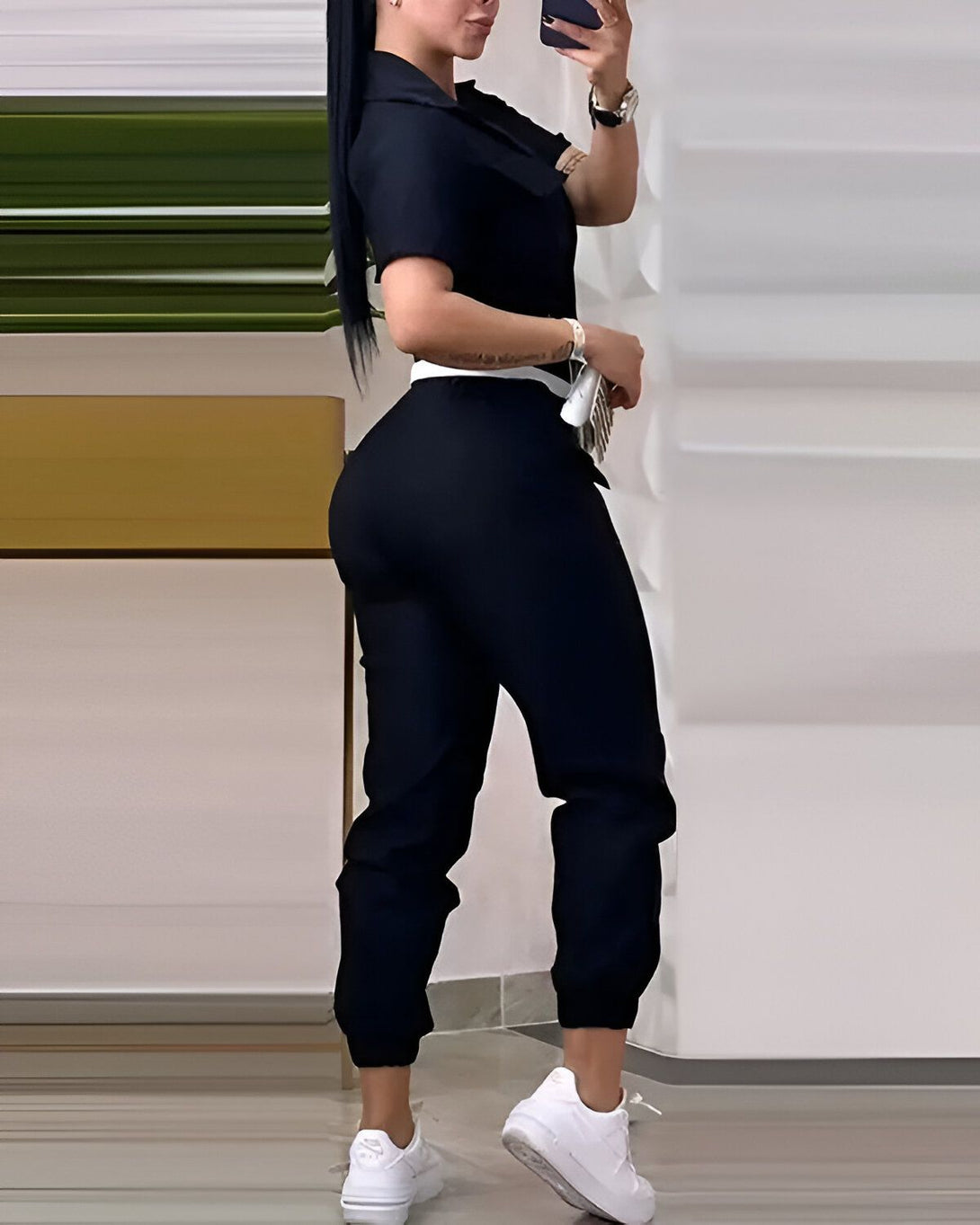 Une femme porte une combinaison noire avec des baskets blanches. Elle est vue de dos et tient son téléphone à la main.
