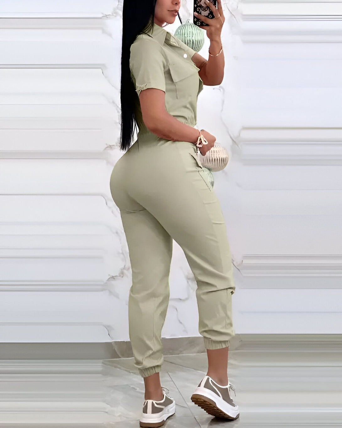Une femme porte une combinaison-pantalon kaki. La combinaison est cintrée à la taille et possède des poches cargo sur les côtés. La femme est vue de dos et porte des baskets blanches.