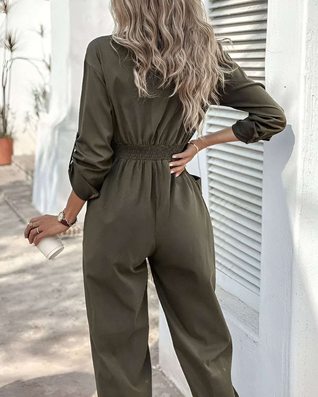 Une femme porte une combinaison-pantalon verte. Elle est vue de dos. La combinaison-pantalon est cintrée à la taille et possède des poches sur les côtés. La femme porte également une montre et tient une tasse à la main.
