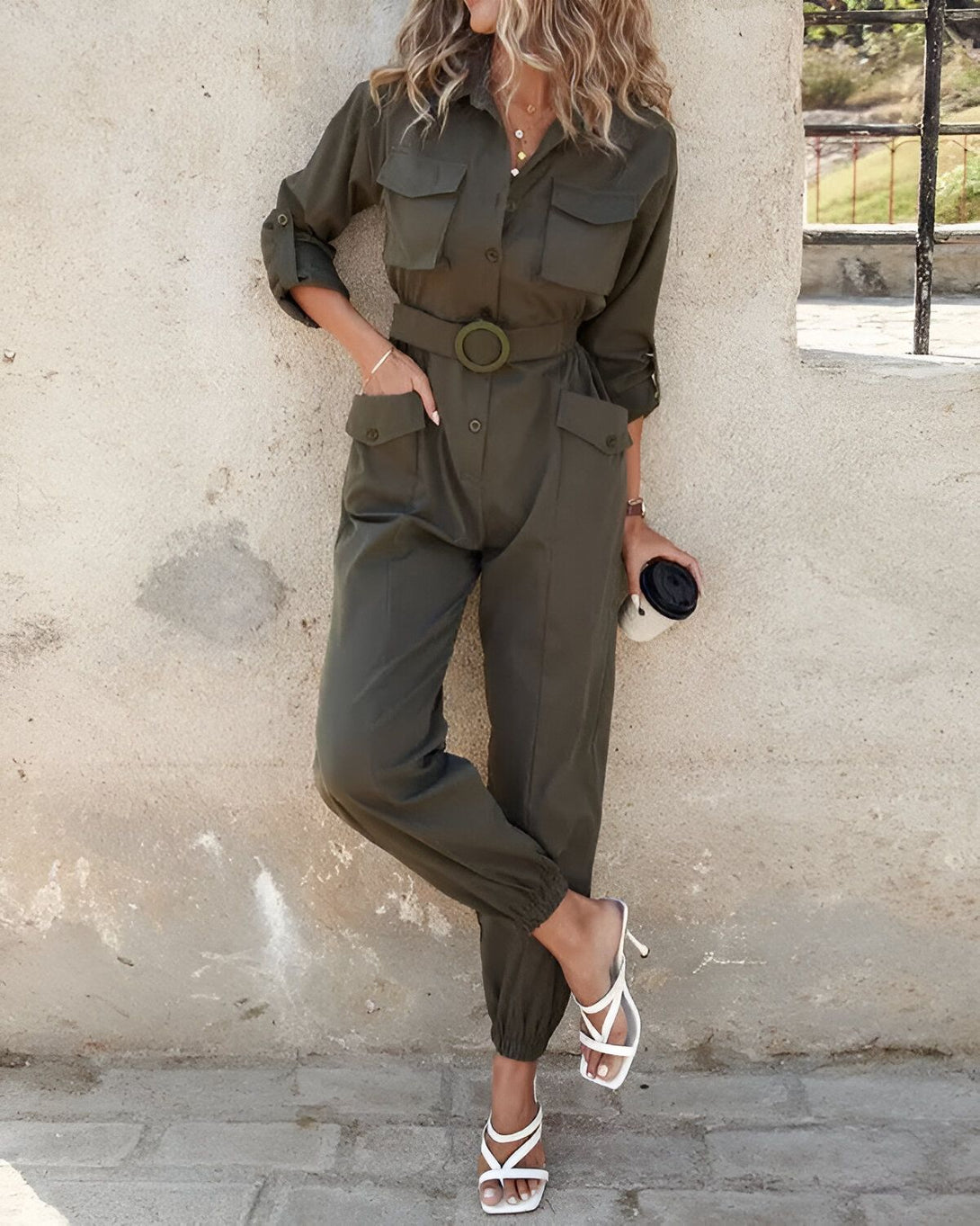 Une femme porte une combinaison-pantalon kaki ceinturée à la taille et des sandales à talons blancs. Elle a une tasse à la main.
