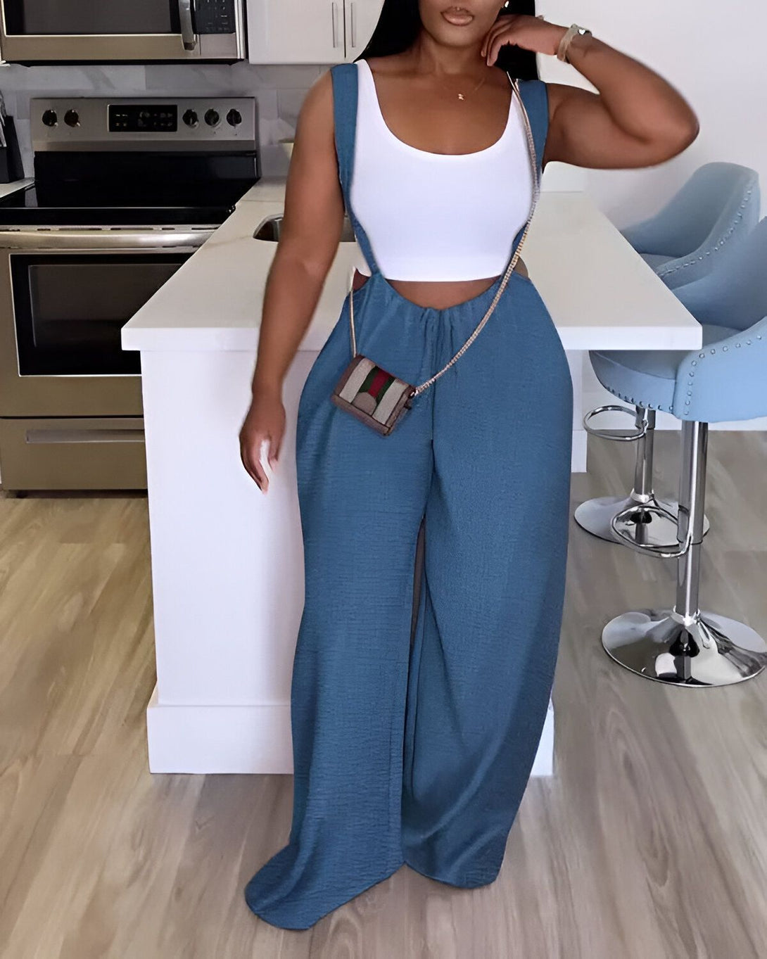 Une femme porte un pantalon large bleu et un haut blanc. Elle est debout dans une cuisine et porte un sac à main marron.