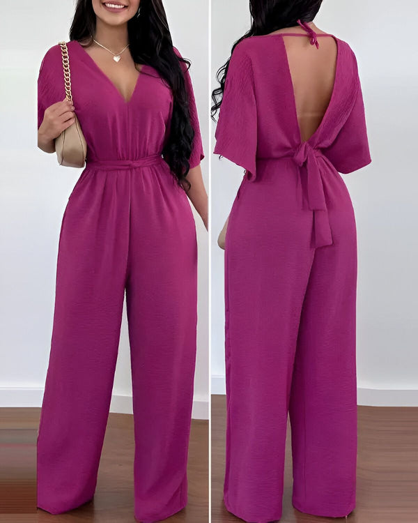 Une femme porte une combinaison-pantalon rose avec un décolleté en V et des manches courtes. La combinaison est cintrée à la taille et possède une ceinture. Le dos de la combinaison est ouvert et noué au niveau de la taille. La femme porte également un collier et un sac à main.