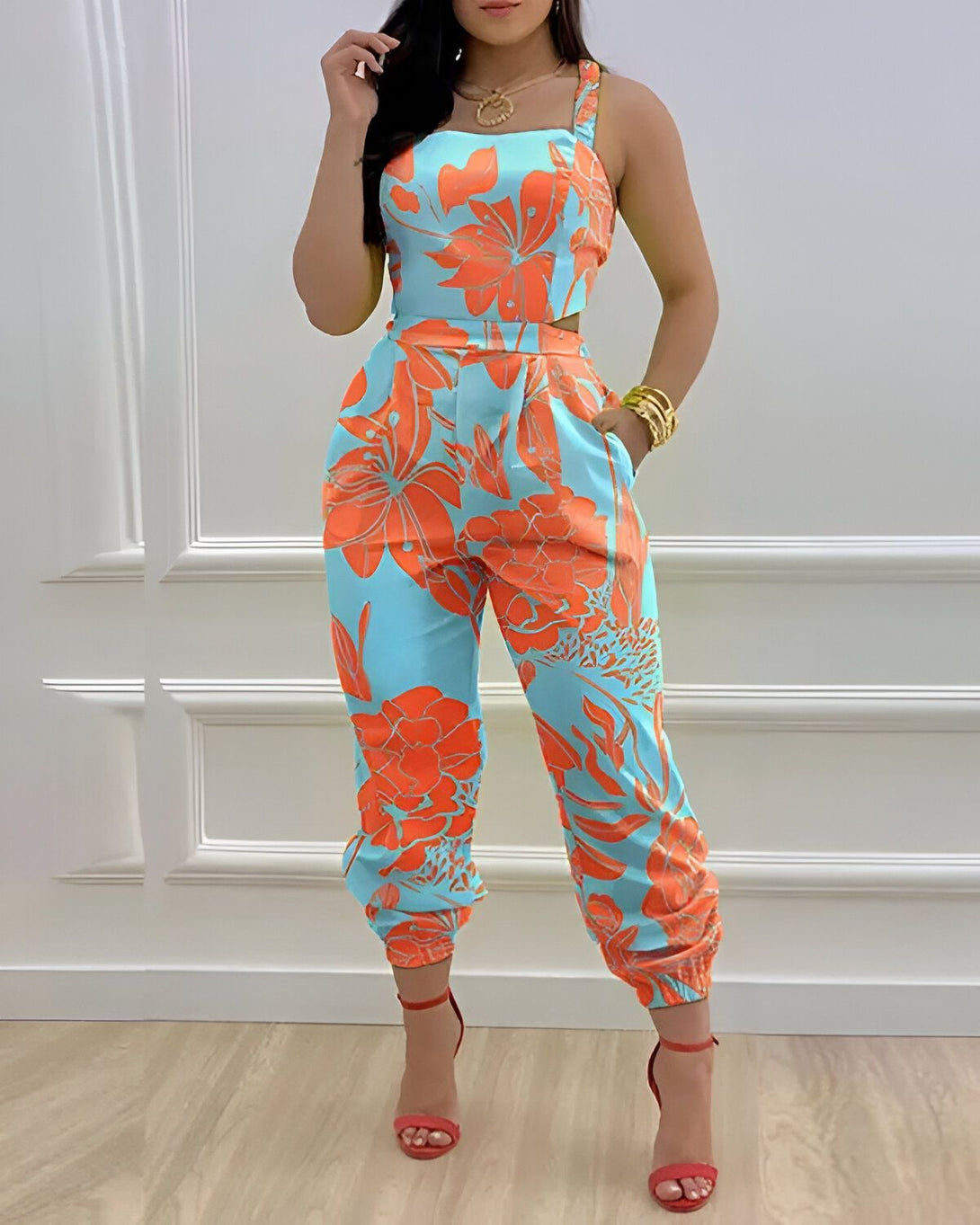 Une femme porte une combinaison pantalon orange et bleu à motifs floraux. Elle est assortie de sandales à talons hauts rouges et d'un collier doré.