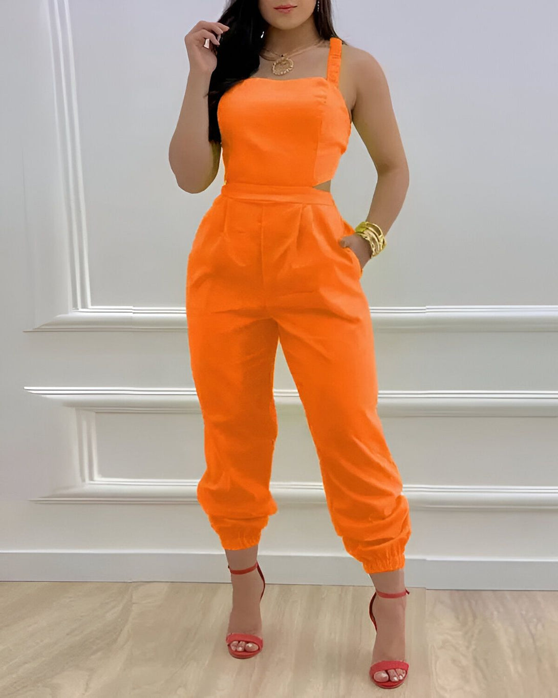 Une femme porte une combinaison orange à bretelles avec des talons hauts orange. Elle est debout dans une pièce blanche avec des murs blancs et un sol en bois.