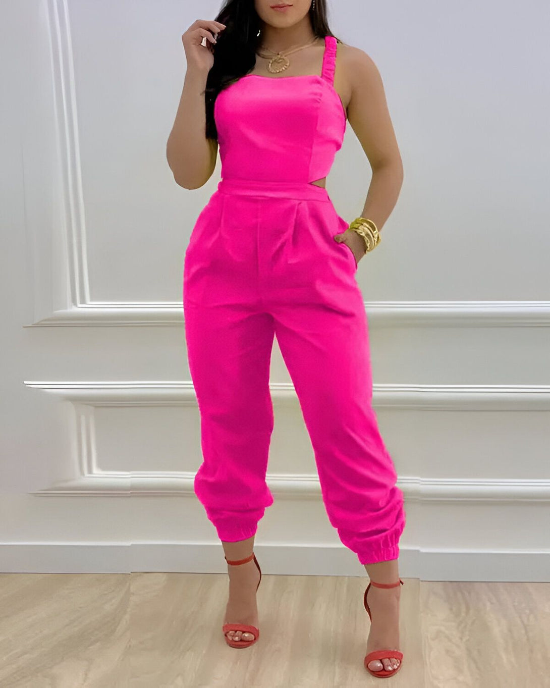 Une femme porte une combinaison pantalon rose fuchsia. La combinaison est près du corps et a une coupe évasée au niveau des jambes. Elle est également dotée d'une encolure carrée et de fines bretelles. La femme porte des sandales à talons rouges et des bijoux dorés. Elle a les cheveux longs et noirs et un maquillage léger. Elle est debout dans un intérieur blanc et pose pour la photo.