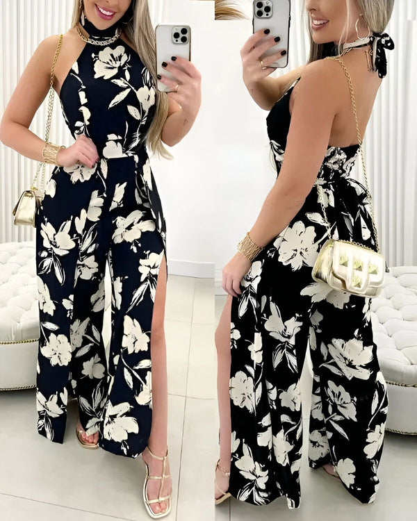 Une femme porte une combinaison pantalon noire et blanche à fleurs. La combinaison a un col montant et des jambes fendues. La femme porte également des sandales à talons noires et un sac à main doré.