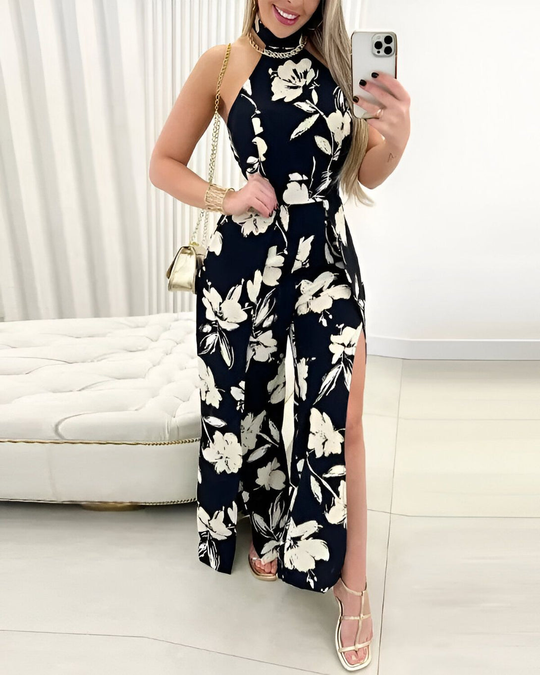Une femme porte une combinaison-pantalon noire à motifs floraux blancs. Elle est dos nu et possède un col montant. La combinaison est fendue sur les côtés. La femme est debout, elle porte des sandales à talons dorées et tient un sac à main doré.