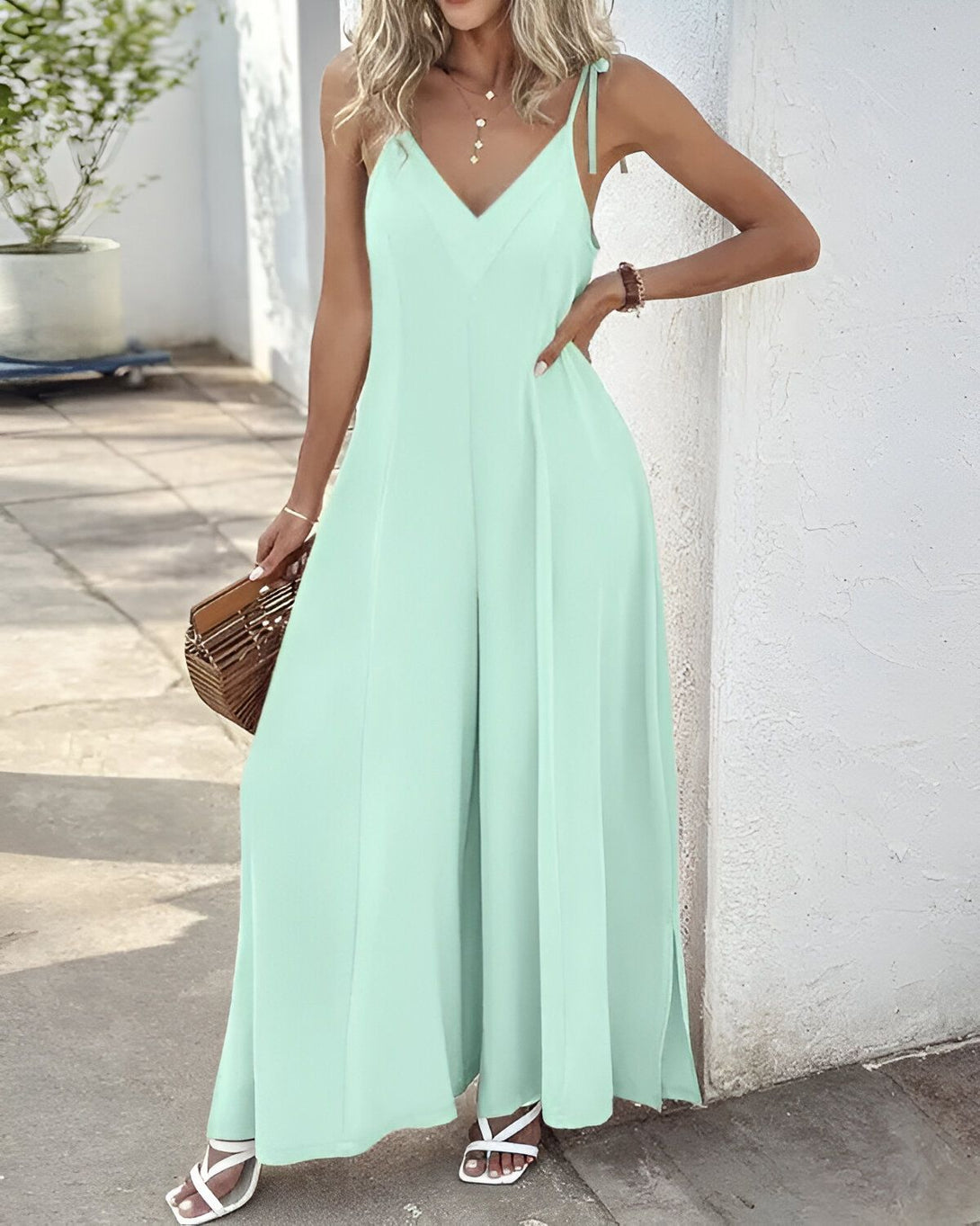 Une femme porte une combinaison-pantalon vert menthe à bretelles. Elle est fendue sur le côté et possède une encolure en V. La femme est blonde et porte des sandales blanches. Elle a un sac à main marron.