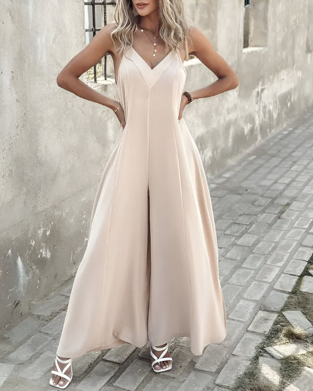 Une femme porte une combinaison-pantalon beige. Elle est debout, les mains sur les hanches. La combinaison-pantalon est ample et a des jambes larges. Elle porte des sandales blanches à talons hauts.